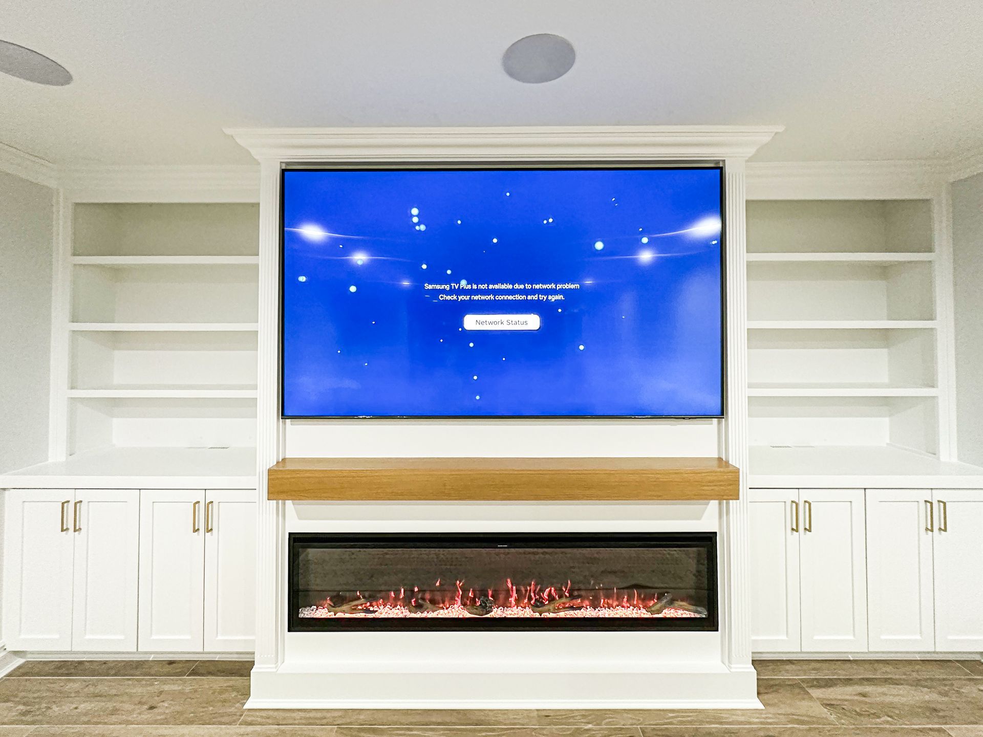 A wall-mounted TV over a linear electric fireplace, flanked by white built-in shelving and cabinets in a living room.
