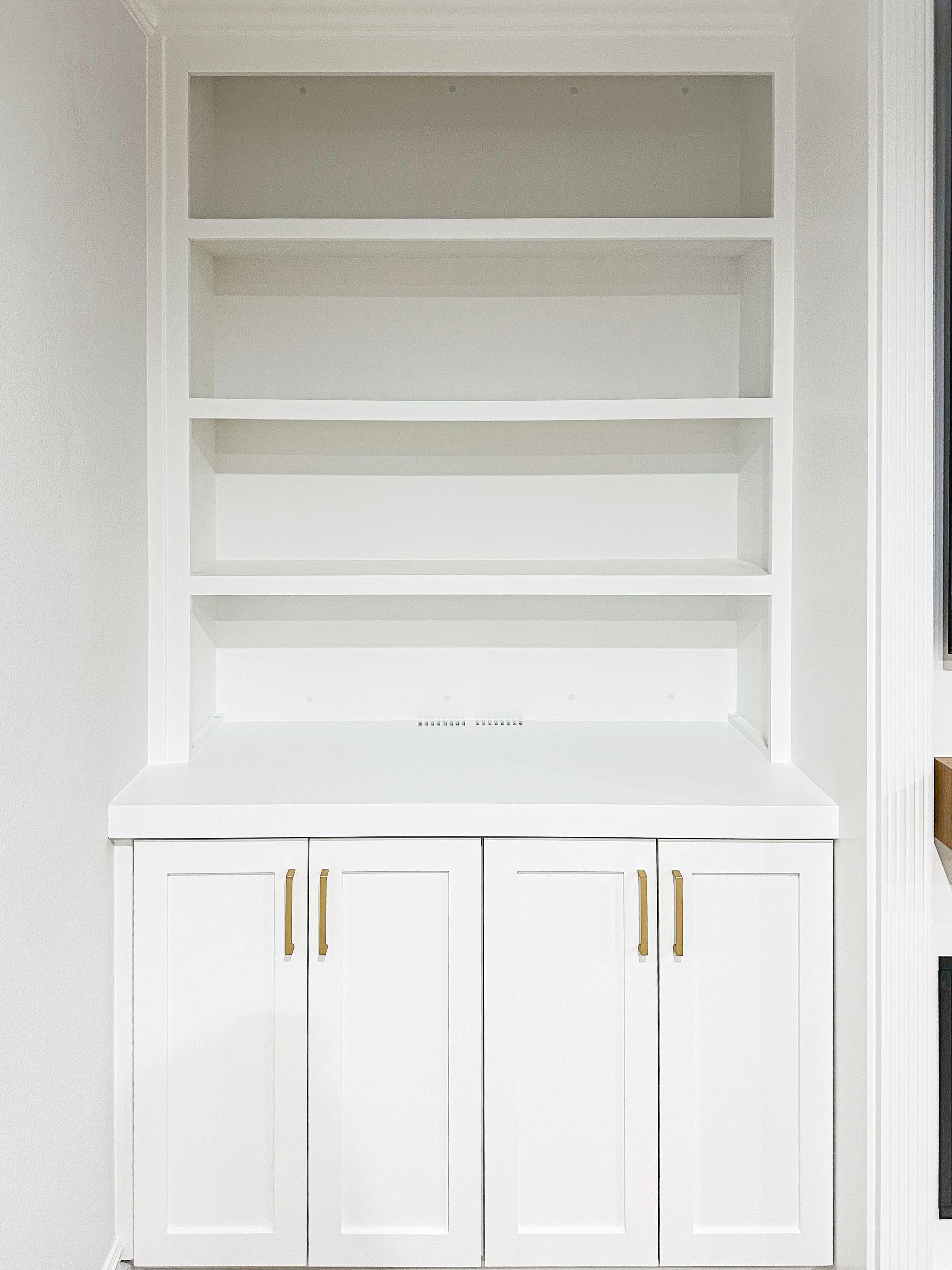 A white, built-in shelving unit with four open shelves above a cabinet base featuring brass-colored vertical handles.
