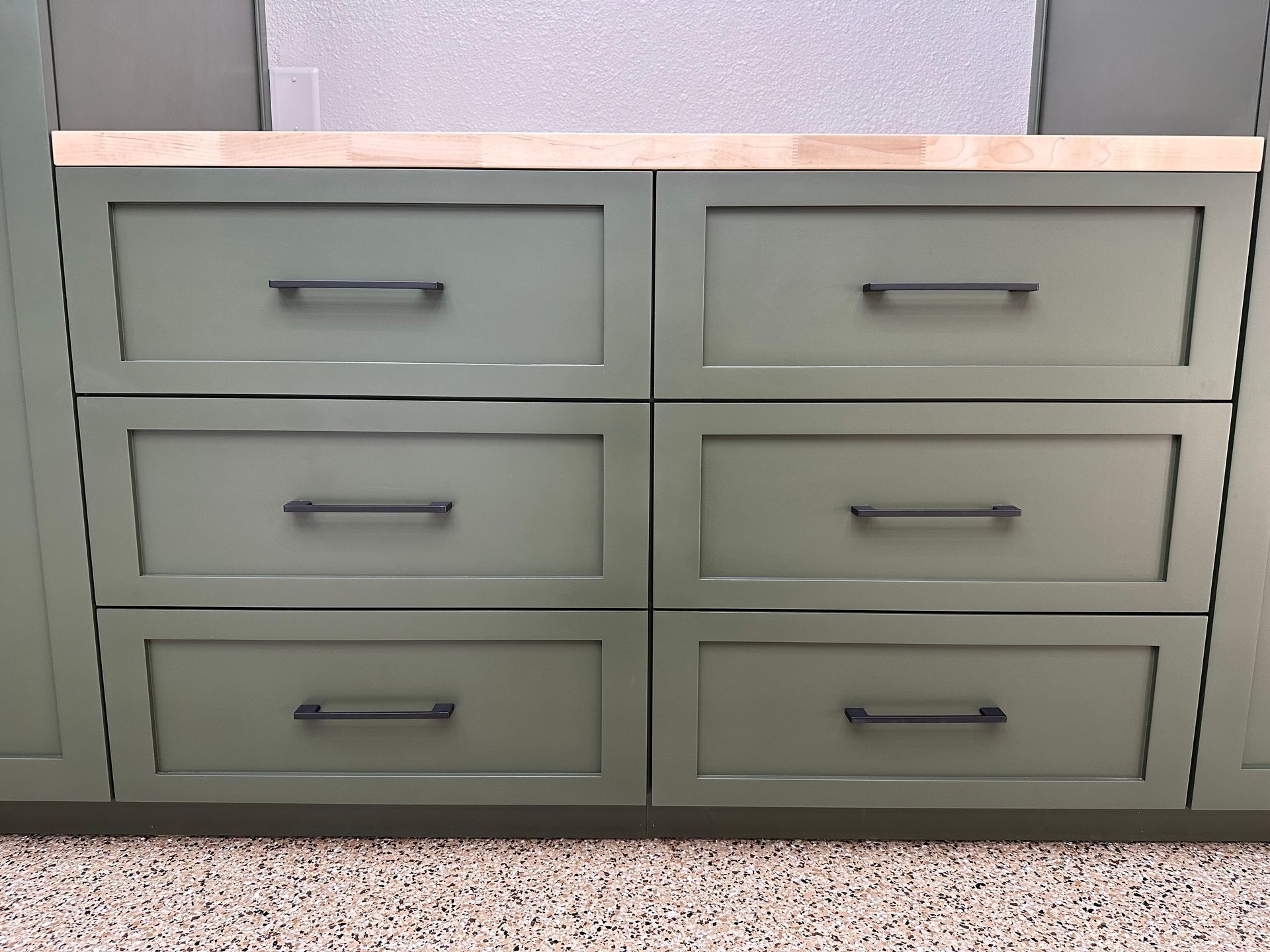 A sage green dresser with six drawers and matte black bar handles, topped with a light wood counter.