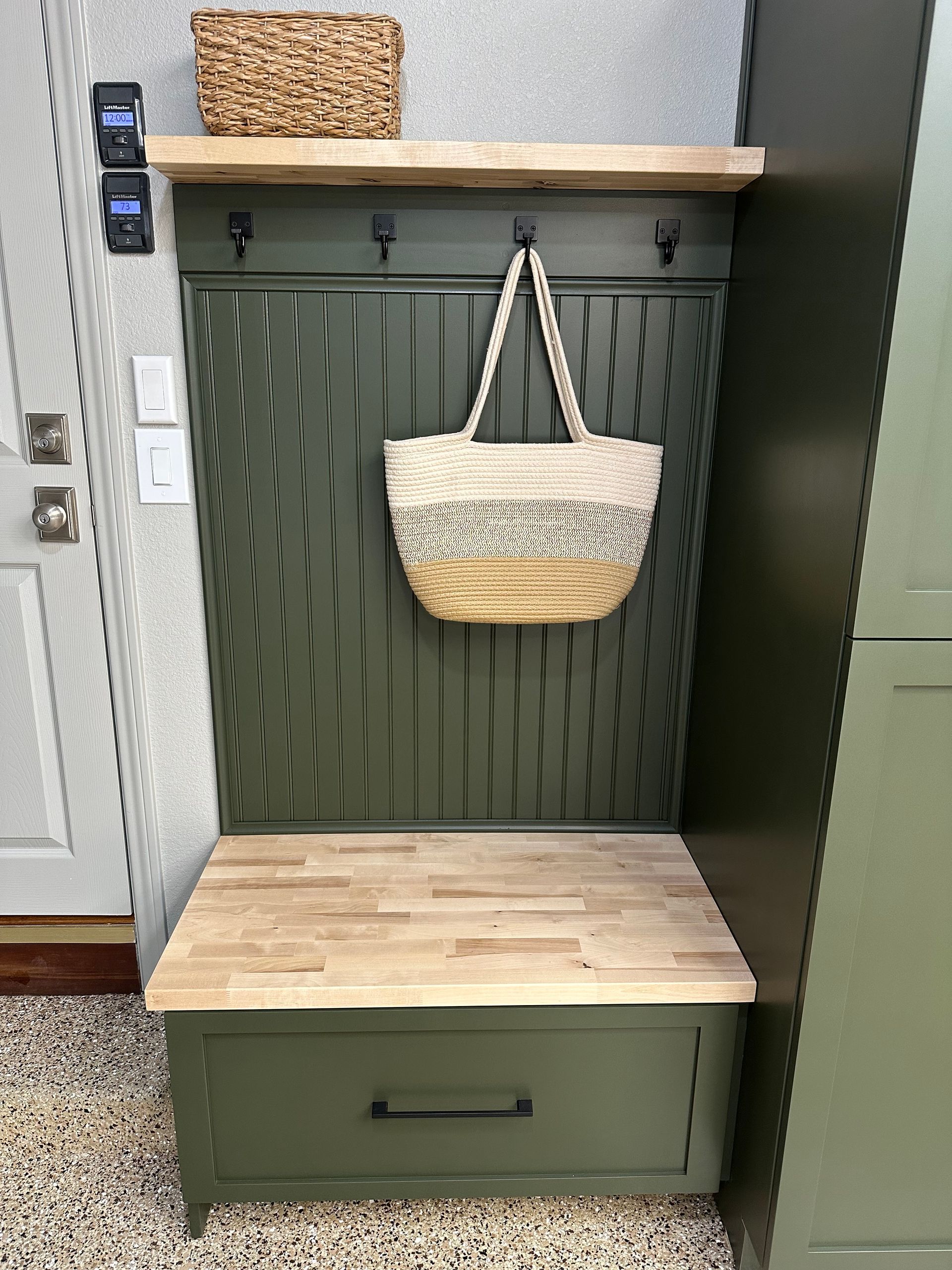 A dark green entryway bench with a light wood seat, storage drawer, a shelf with a wicker basket, and hanging hooks.