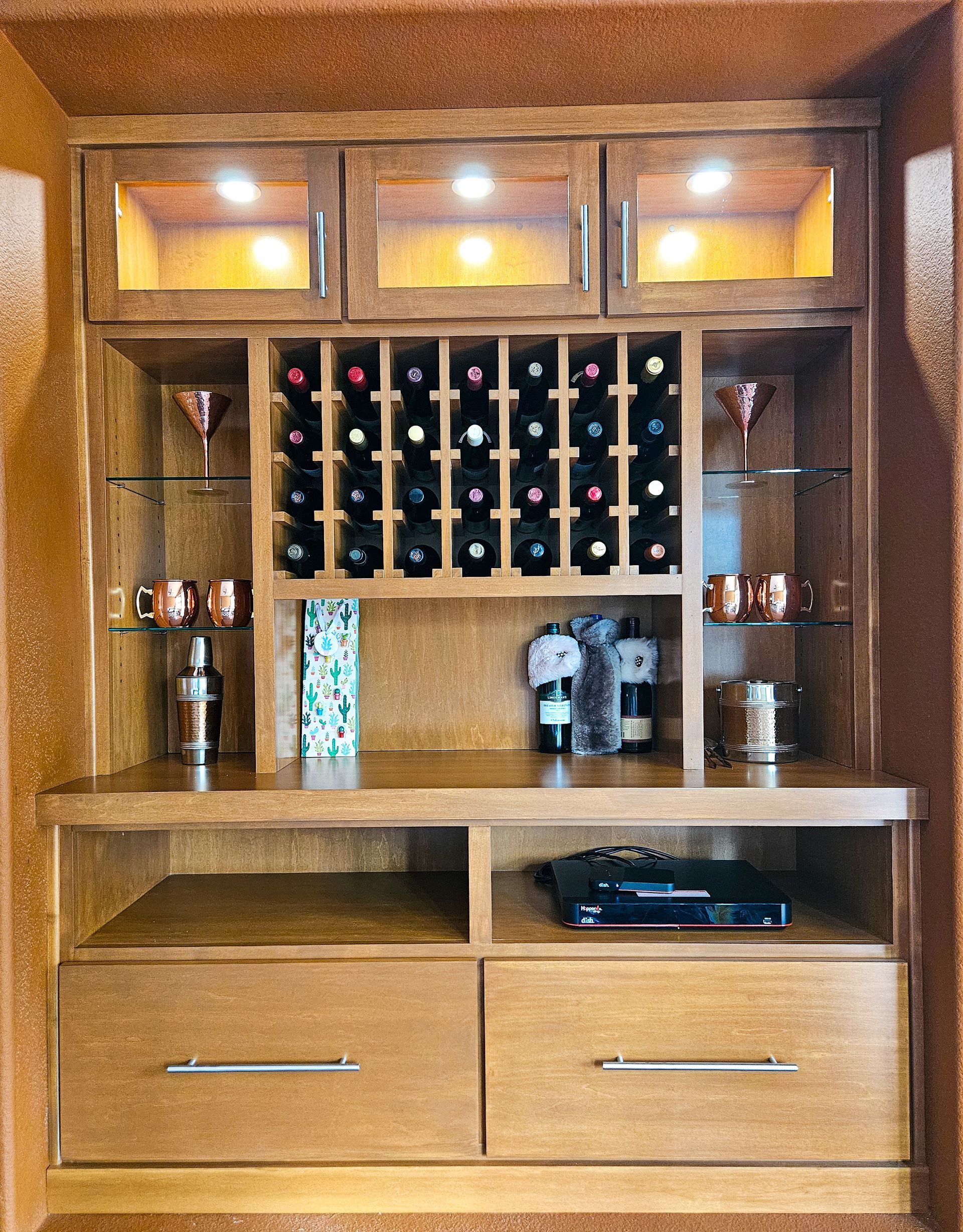A wooden home bar unit with overhead glass-front cabinets, a wine rack, open shelving with glassware, and two bottom drawers.