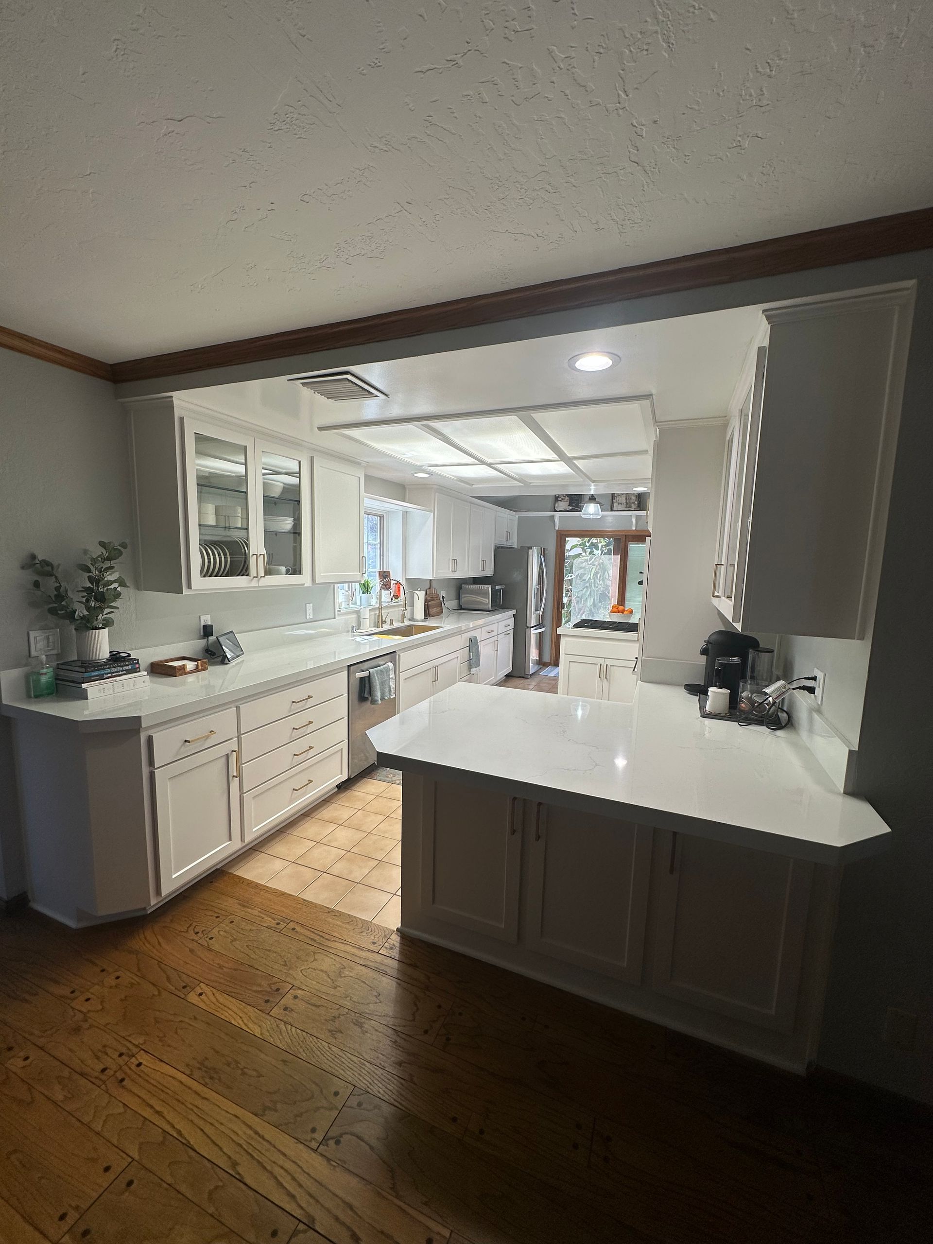 A bright kitchen with white cabinets, light countertops, and wood flooring, viewed from an adjacent room.