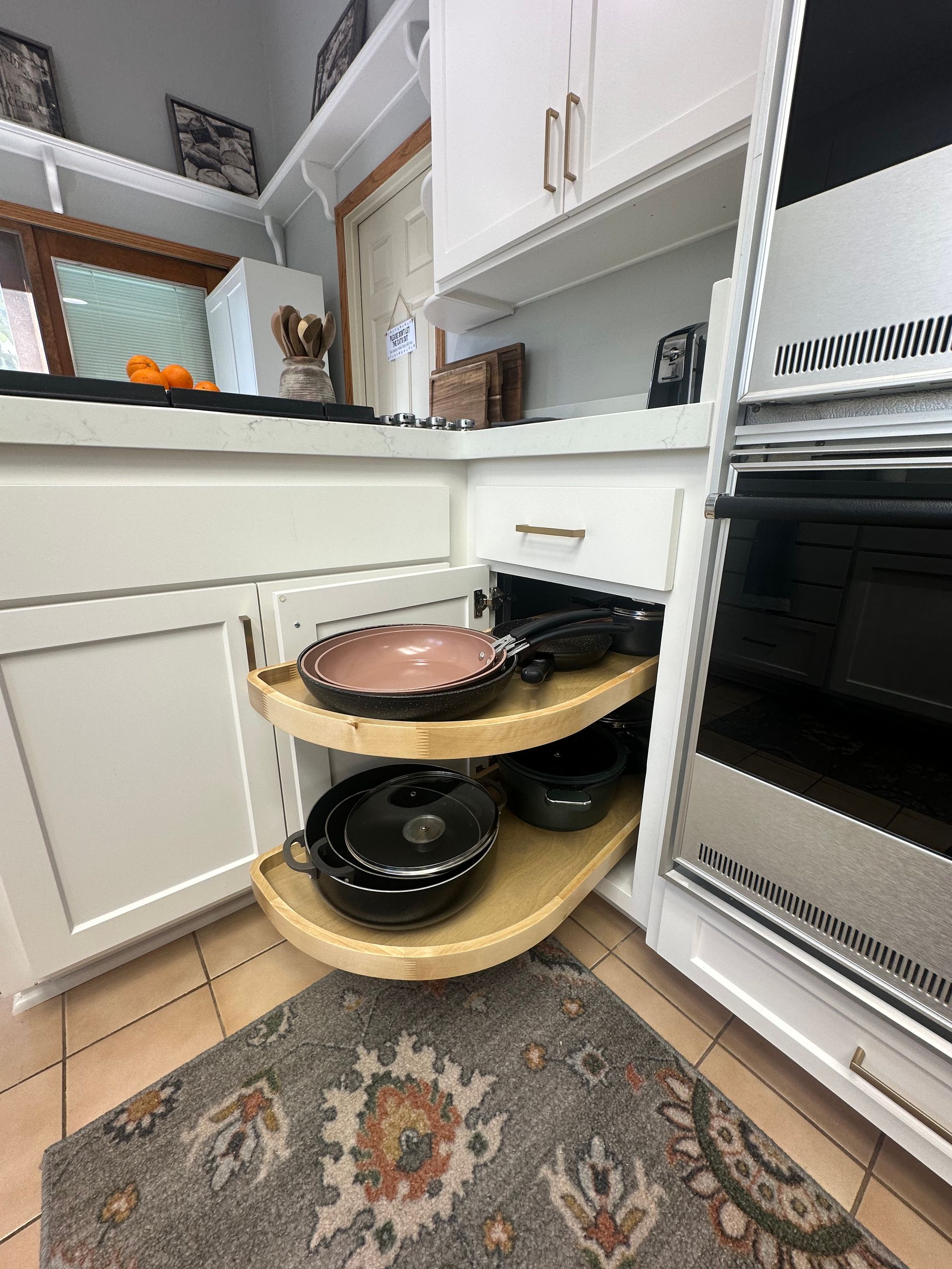 A two-tiered, pull-out wooden corner shelf inside a white kitchen cabinet, holding a pink skillet and black cookware.