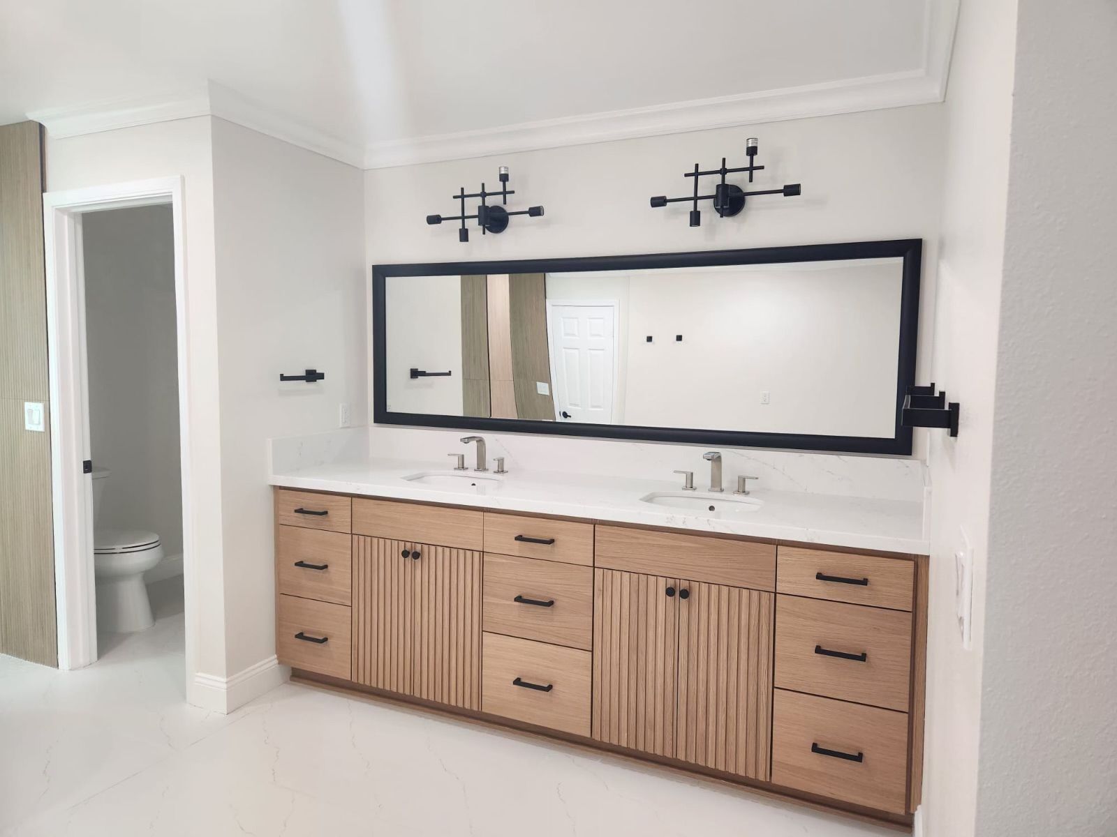 A modern double-sink bathroom vanity with light wood cabinets, black hardware, a large mirror, and industrial lighting.