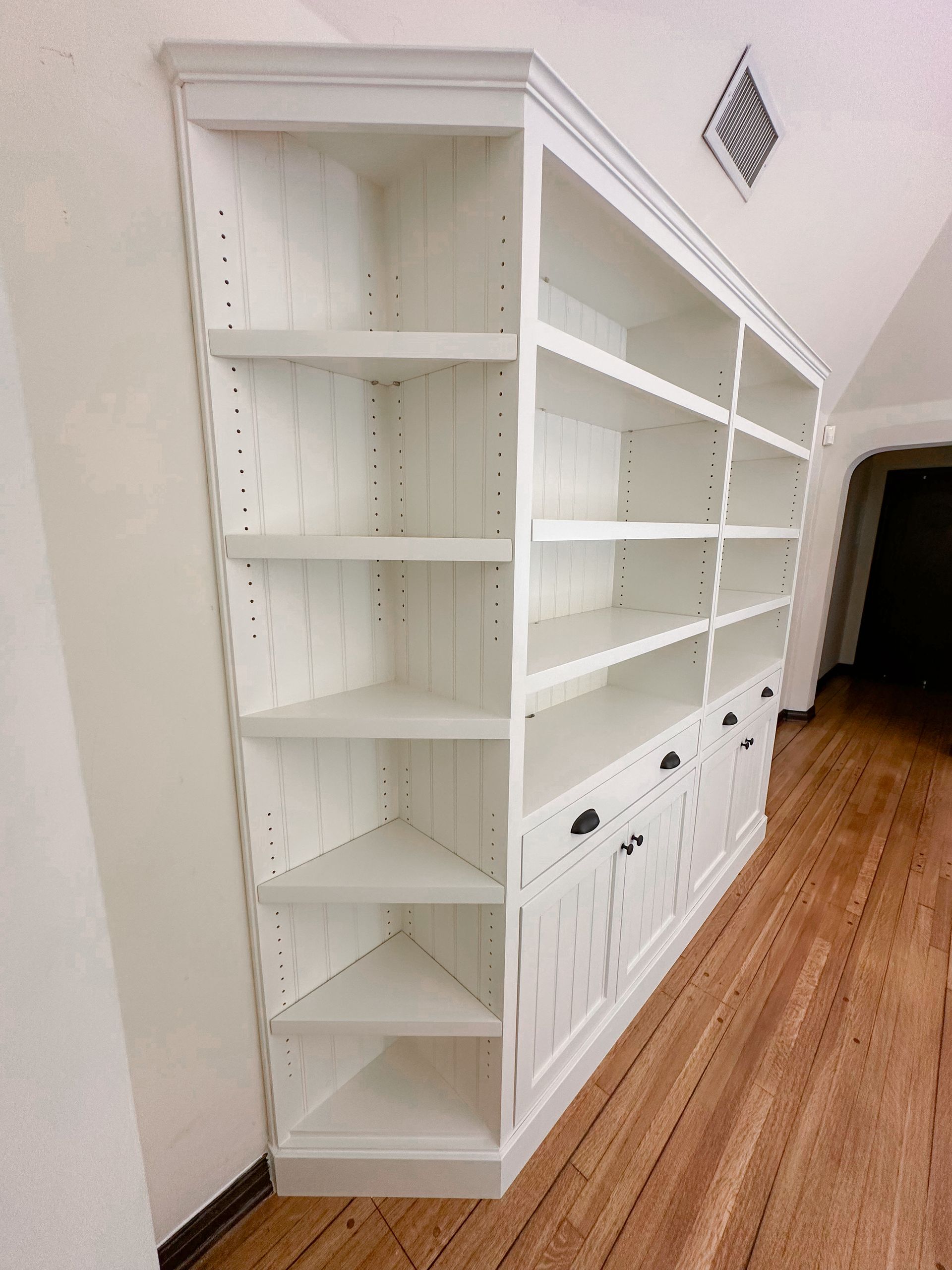 A white, multi-section wooden bookshelf with base cabinets and open shelves, positioned against a wall on a wooden floor.