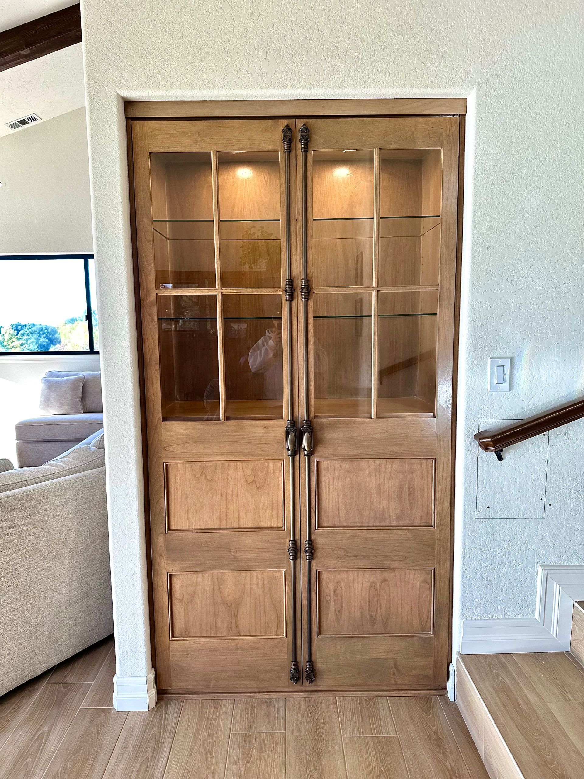 A built-in wooden display cabinet with glass-paned upper doors, lower cabinet panels, and vertical bronze hardware.
