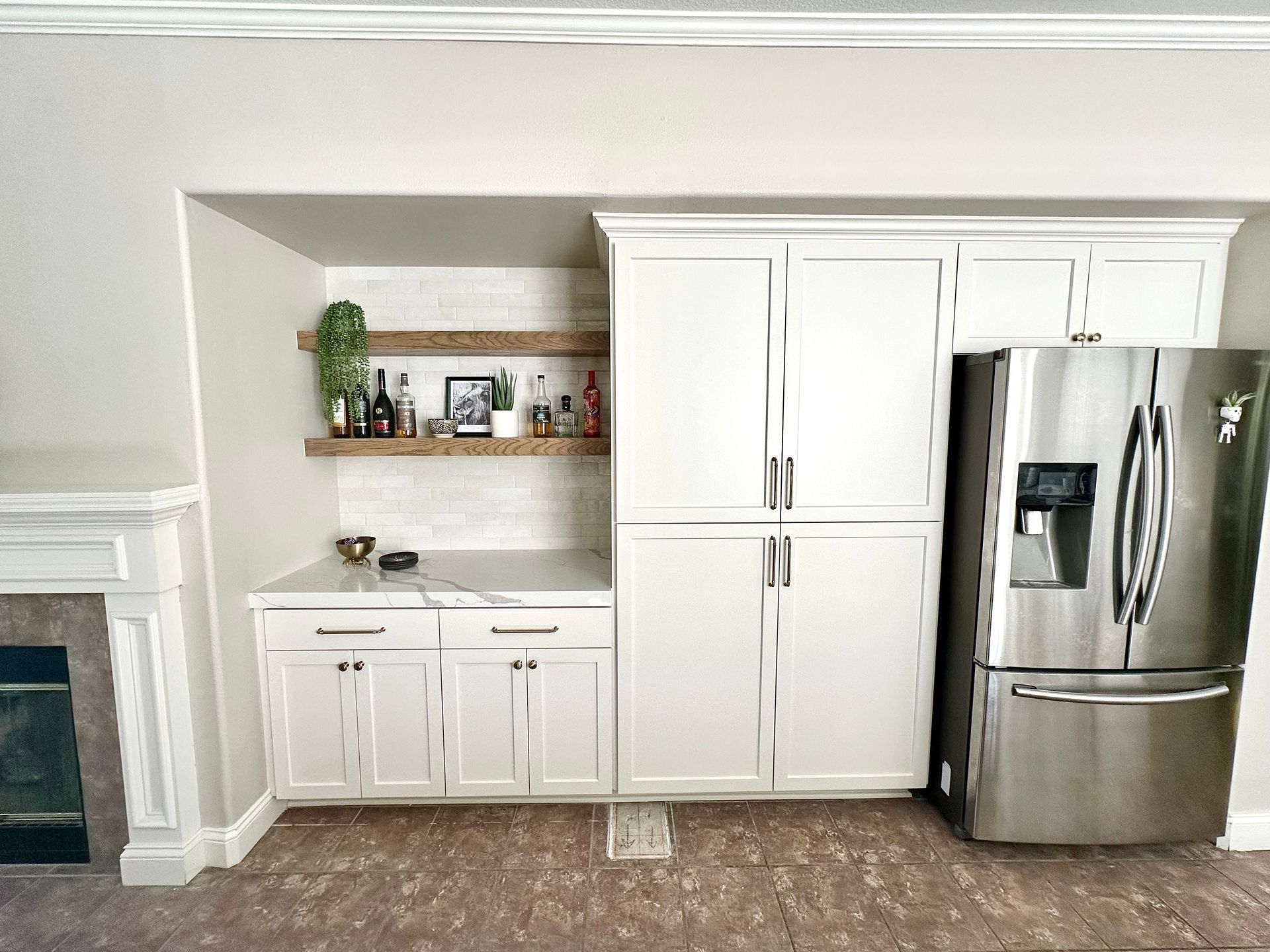 A kitchen wet bar with white cabinets, wood shelves, and a stainless steel refrigerator next to a fireplace.