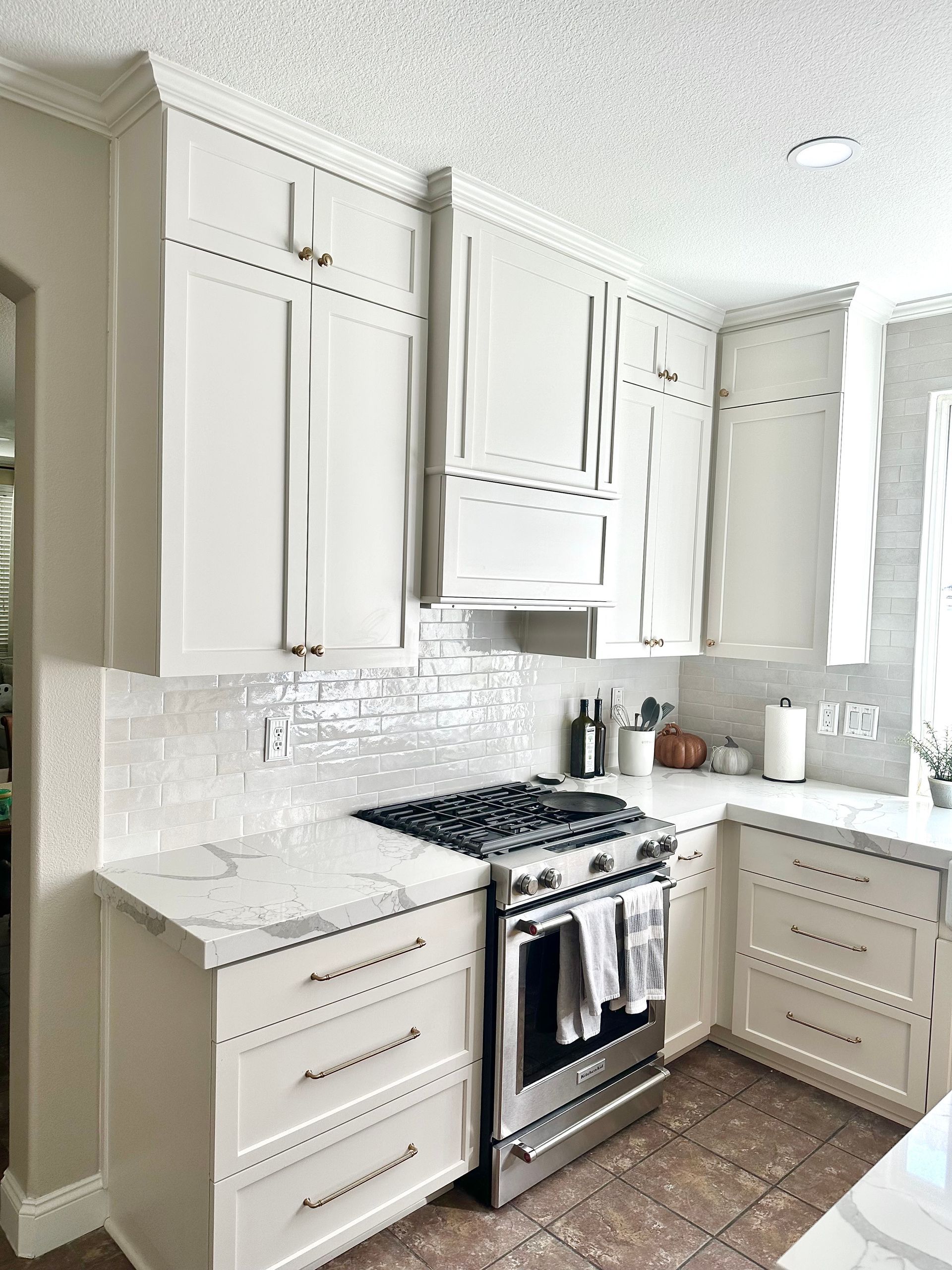 A modern kitchen with off-white cabinets, a stainless steel gas range, marble-patterned countertops, and a tiled backsplash.