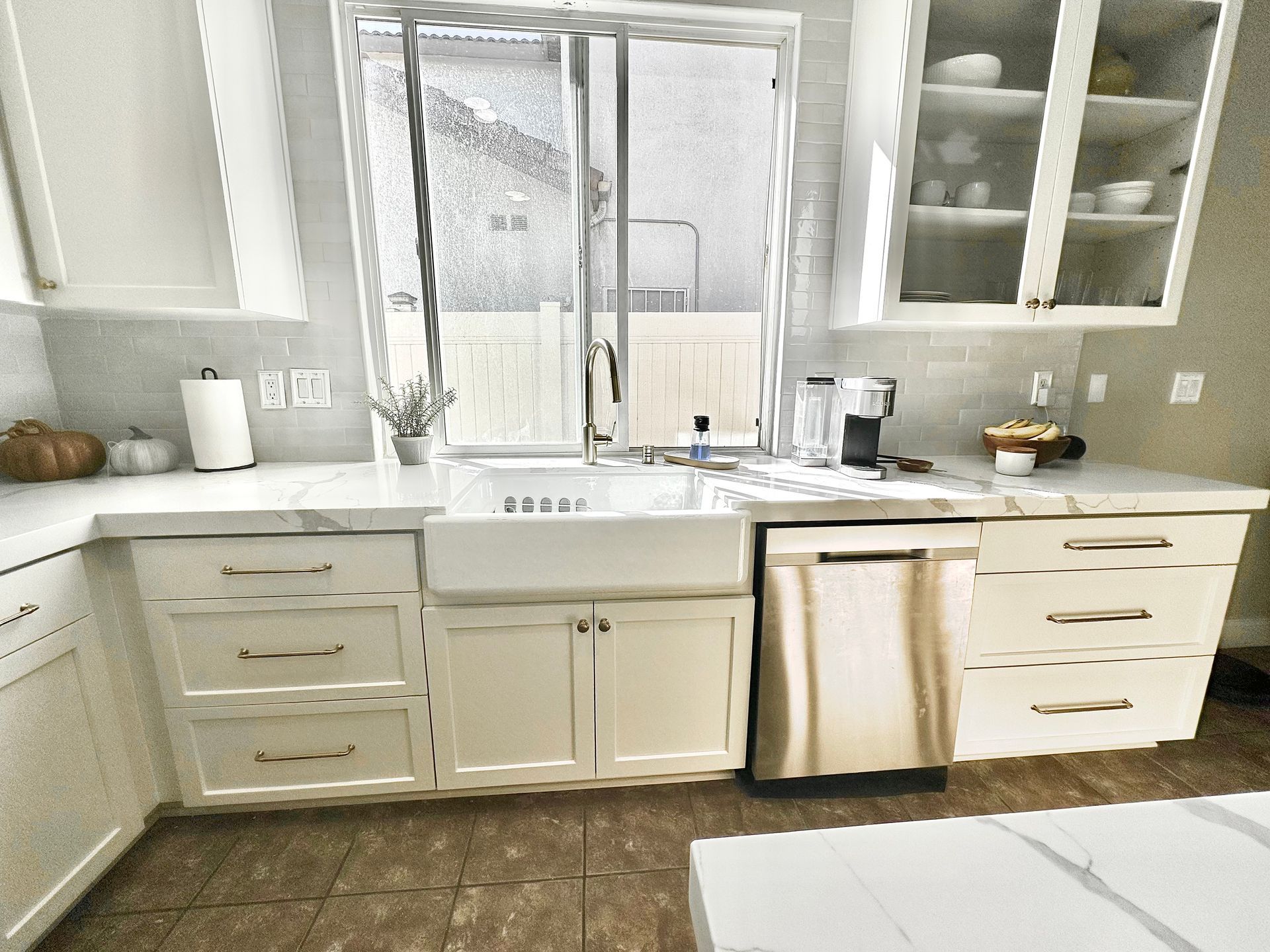 A bright, modern kitchen featuring white cabinets, a farmhouse sink, stainless steel dishwasher, and marble countertops.