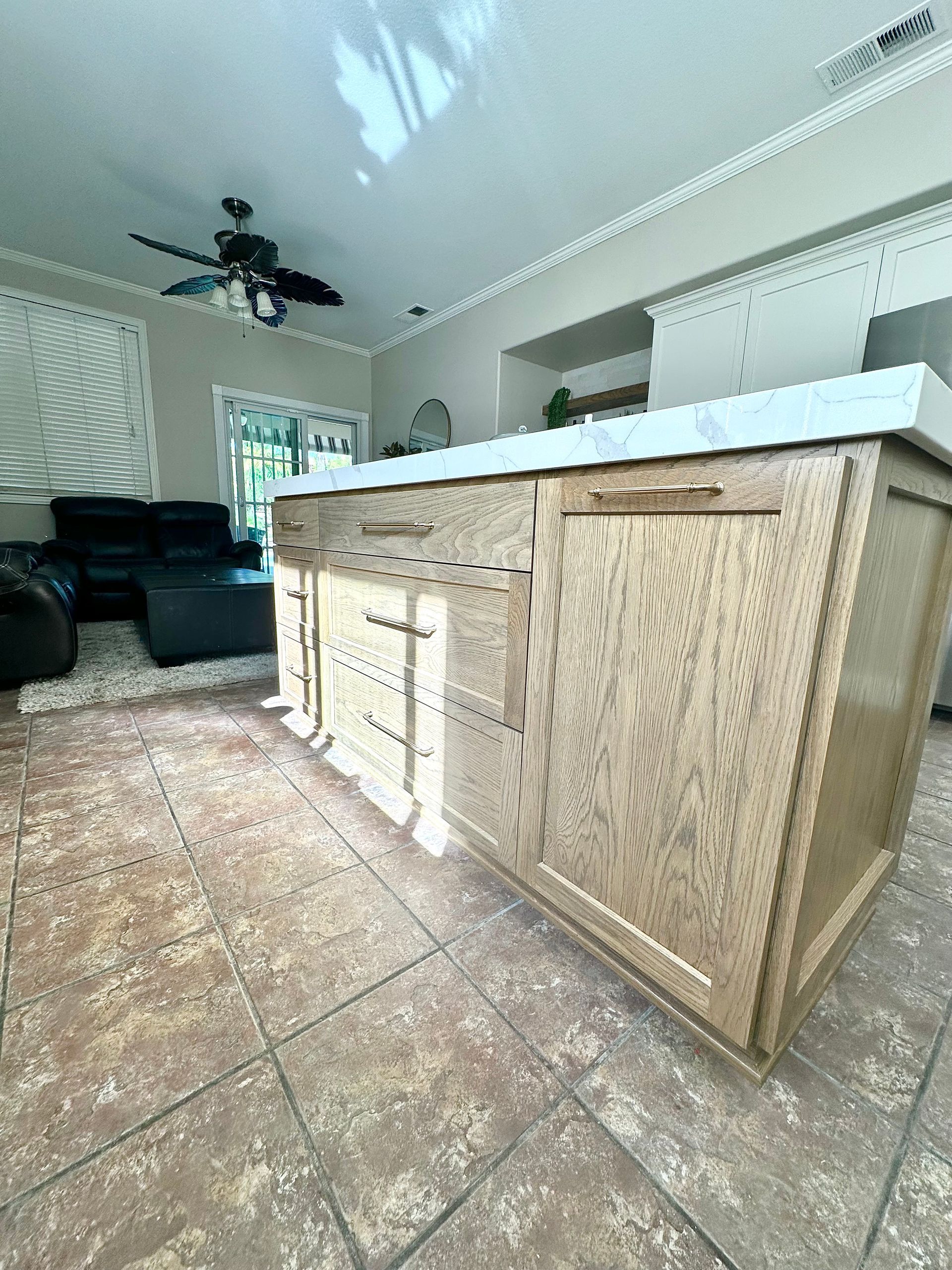 A light-wood kitchen island with a white countertop sits on a tile floor in an open-concept room with a ceiling fan.
