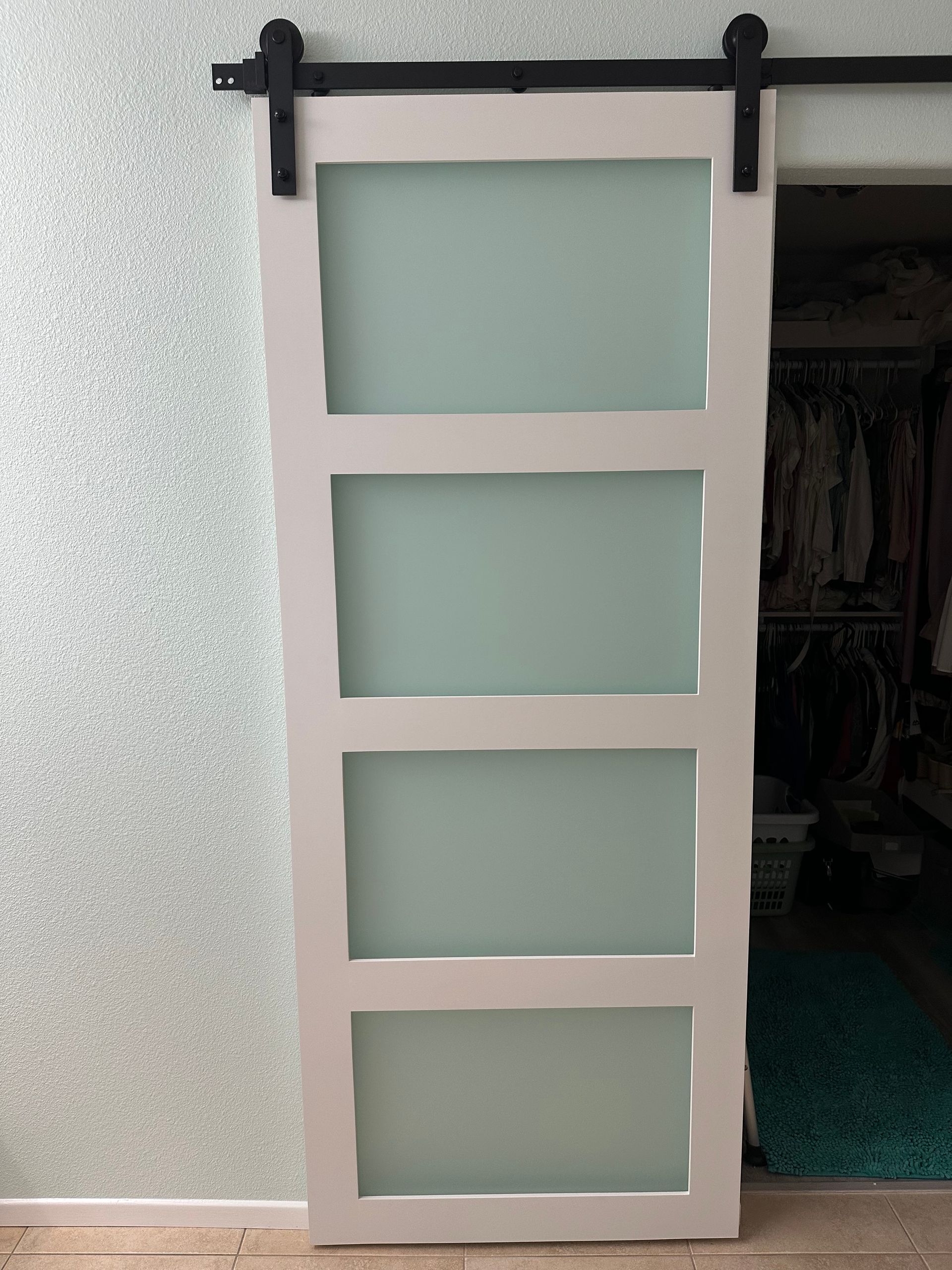 A white, four-panel sliding barn door with frosted glass inserts hangs on a black metal track against a textured wall.
