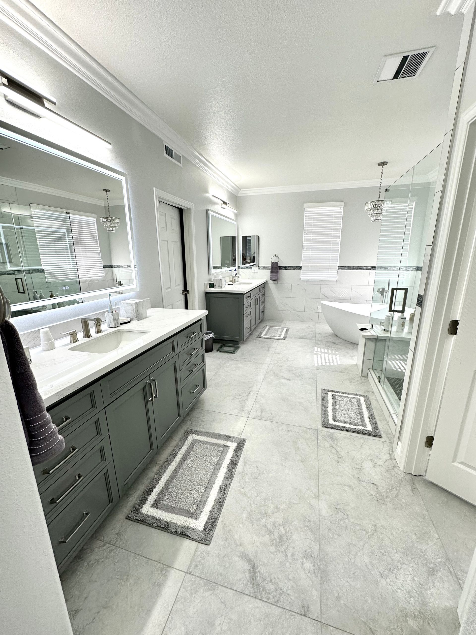 A modern, spacious bathroom with a dark gray double vanity, white marble tile flooring, a soaking tub, and a glass shower.