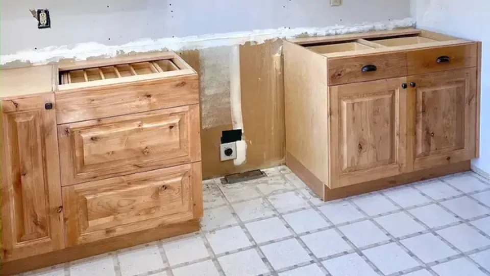 Two wooden kitchen base cabinets installed on a white tiled floor against a wall during a home renovation.