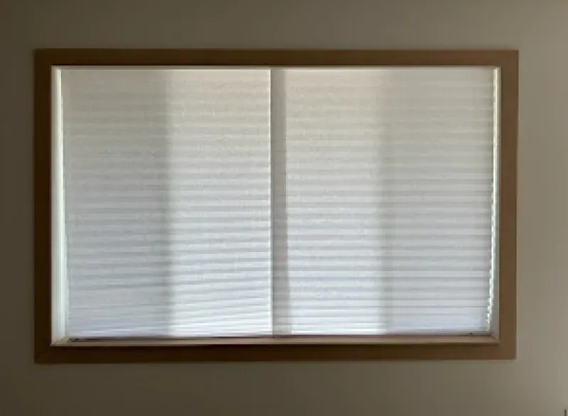 A rectangular window with a light-colored wooden frame, covered by closed, white pleated blinds against a plain wall.