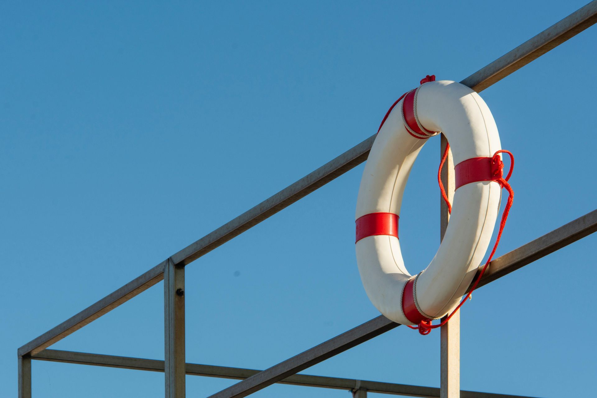 Life preserver ring, white with red stripes, hangs on a metal railing against a clear blue sky.
