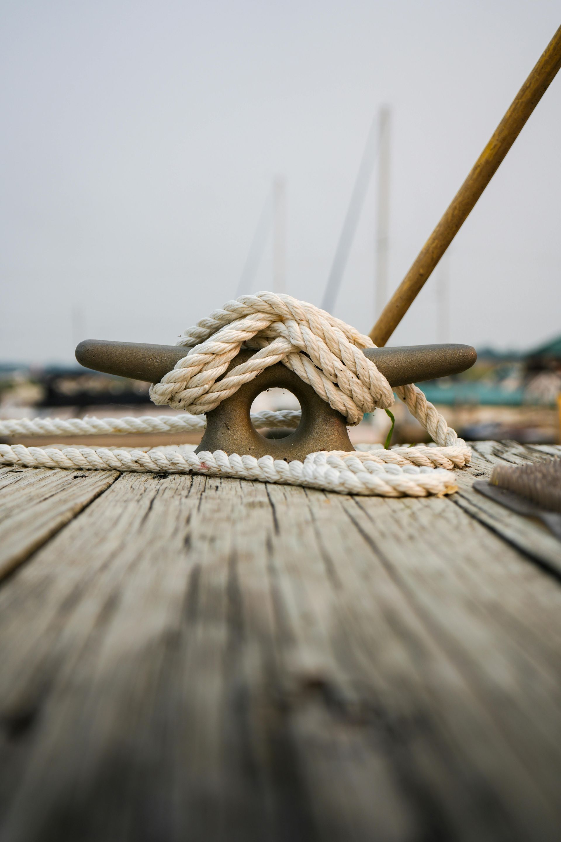 A close-up of a weathered cleat on a wooden dock, with white rope wrapped around it, and sailboats in the background.