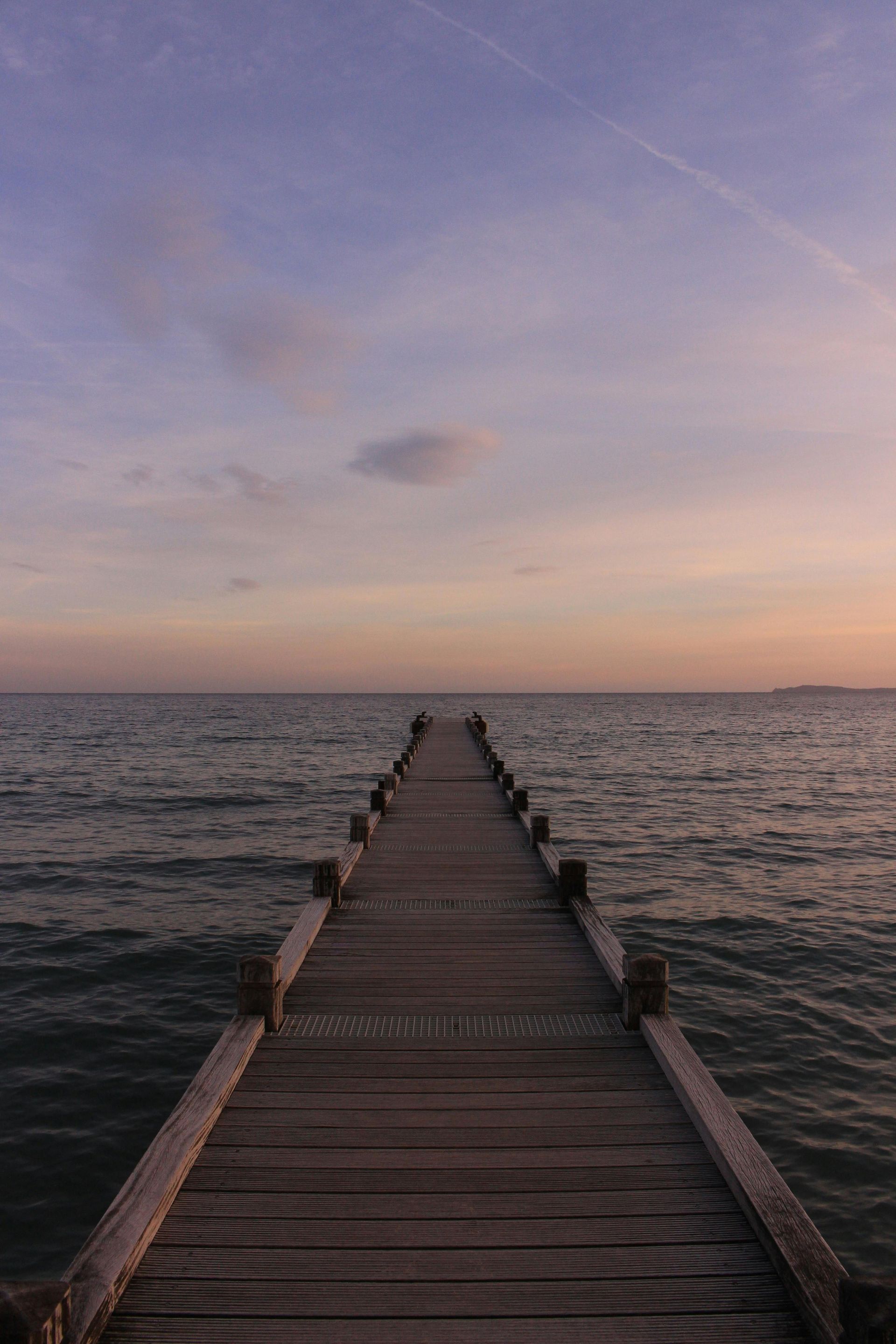 Wooden pier extending towards the horizon over calm water, under a pastel-colored sky at sunset.
