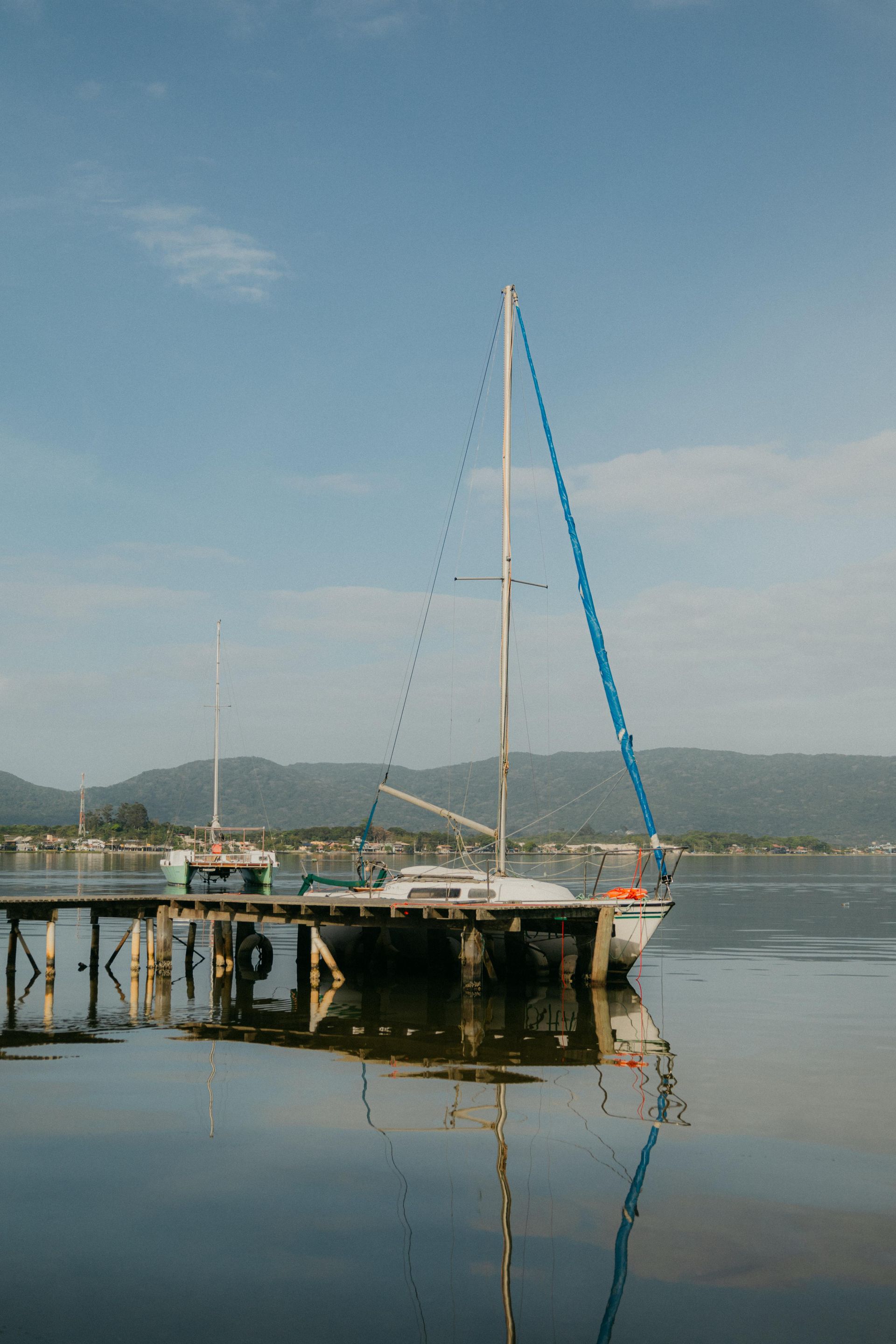 Sailboat docked at a wooden pier, reflecting in still water under a blue sky. Mountains are in the distance.