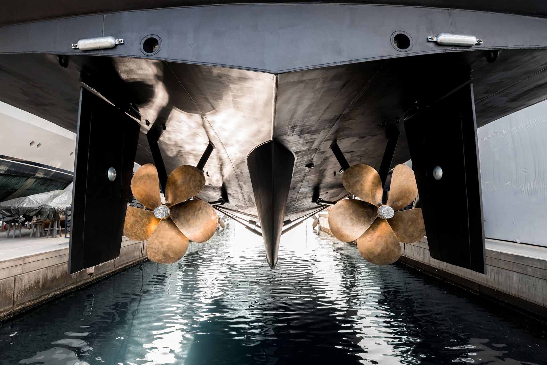 Underside of a large boat showing two propellers in a dock, with the boat's shadow reflecting in the water.