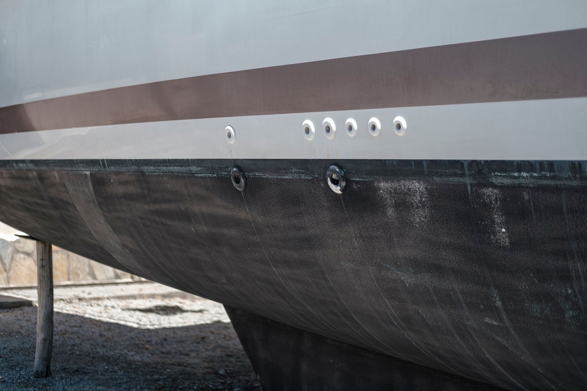 The hull of a boat, showing a series of holes along the side, resting on supports in a drydock.