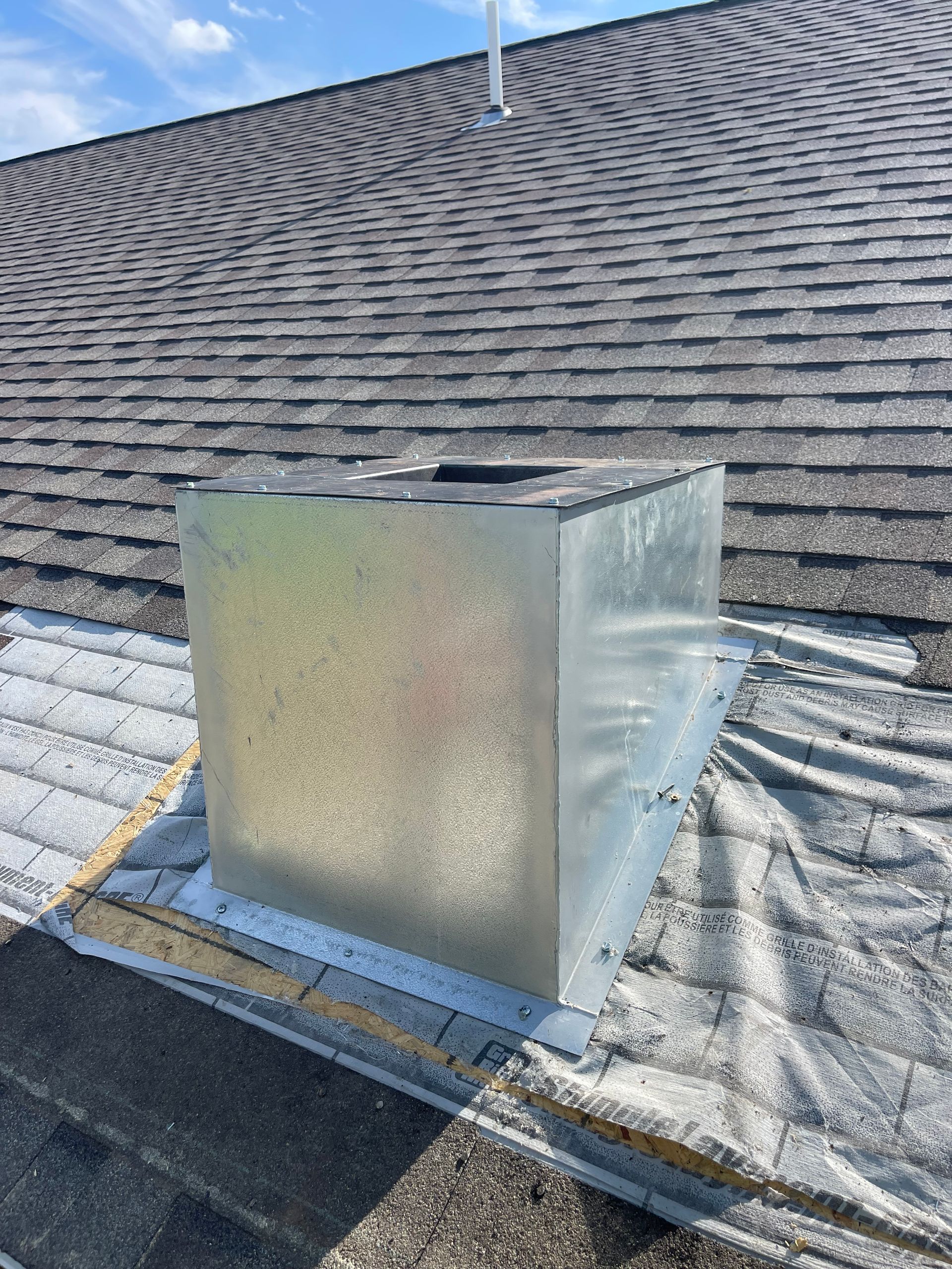 Metal chimney on asphalt shingle roof, surrounded by flashing and a blue sky.