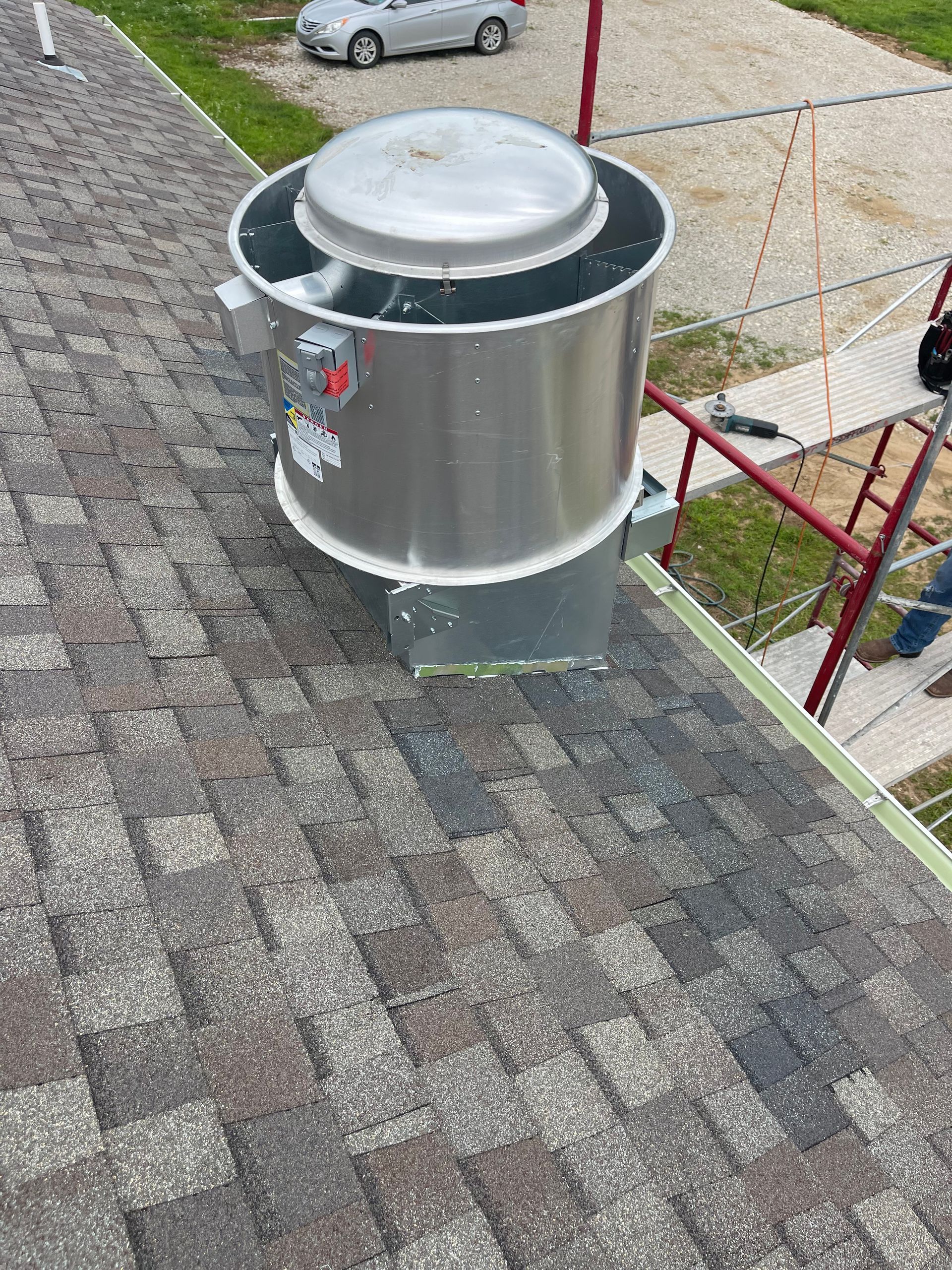 Large cylindrical roof vent on a shingled roof; car and scaffolding in background.