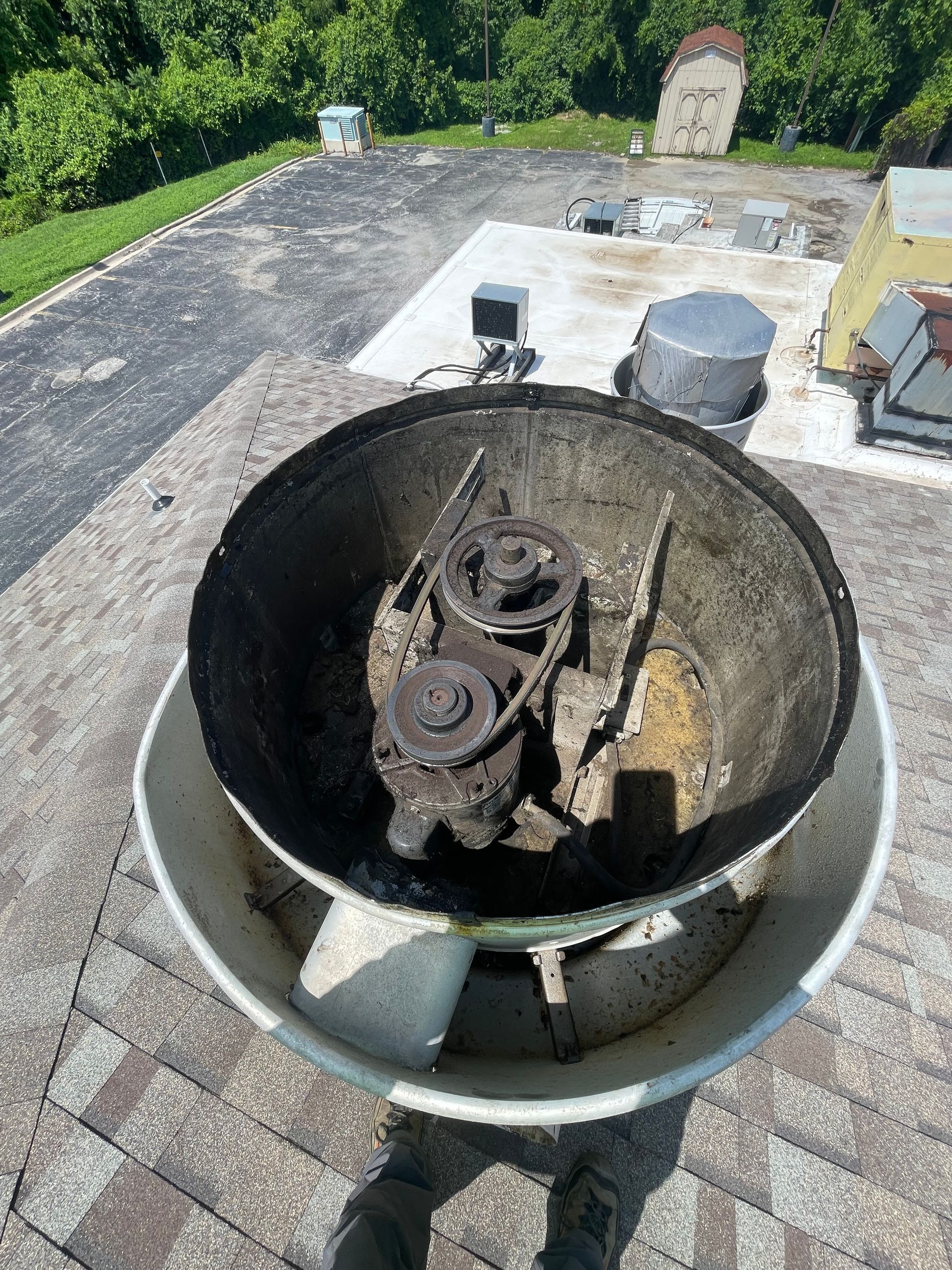 A dirty, open roof-mounted exhaust fan with a complex interior on a rooftop with foliage in the background.