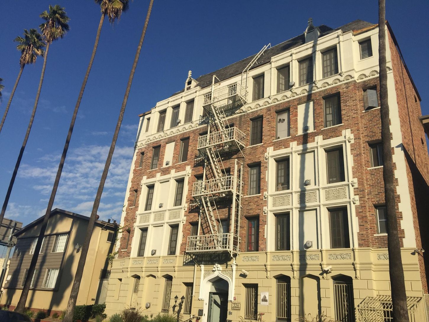 Apartment building with fire escape, brick facade, and palm trees against a blue sky.