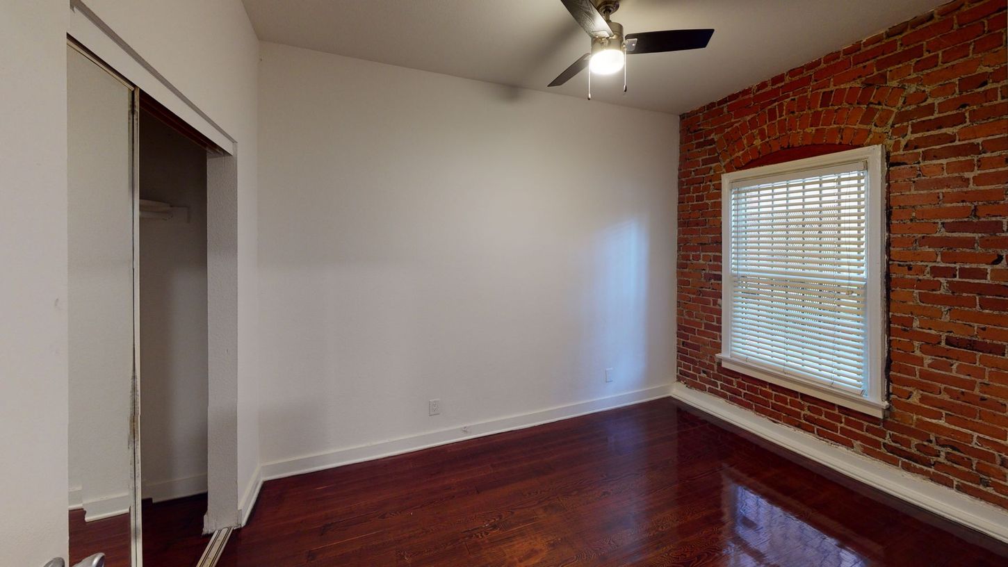 Empty room with hardwood floors, exposed brick wall, window with blinds, and closet.