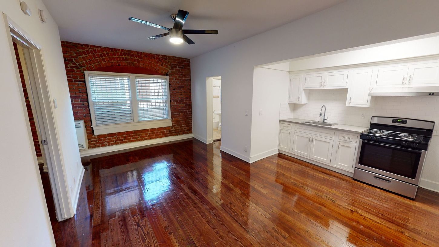 Interior view of an apartment with hardwood floors, exposed brick wall, and kitchen with a stainless steel range.