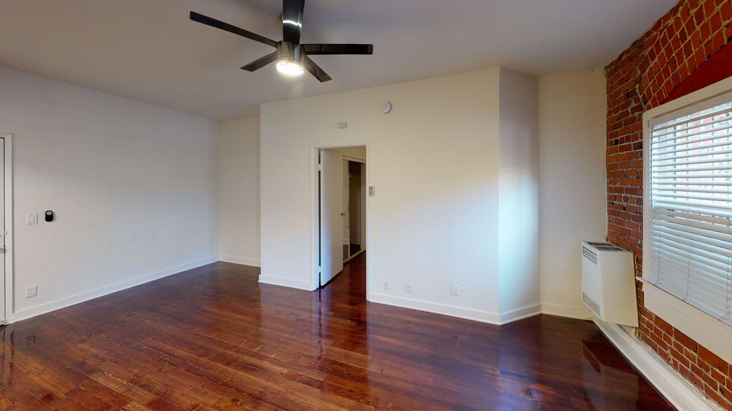 Empty room with dark hardwood floors, white walls, and exposed brick. A ceiling fan and a window are visible.