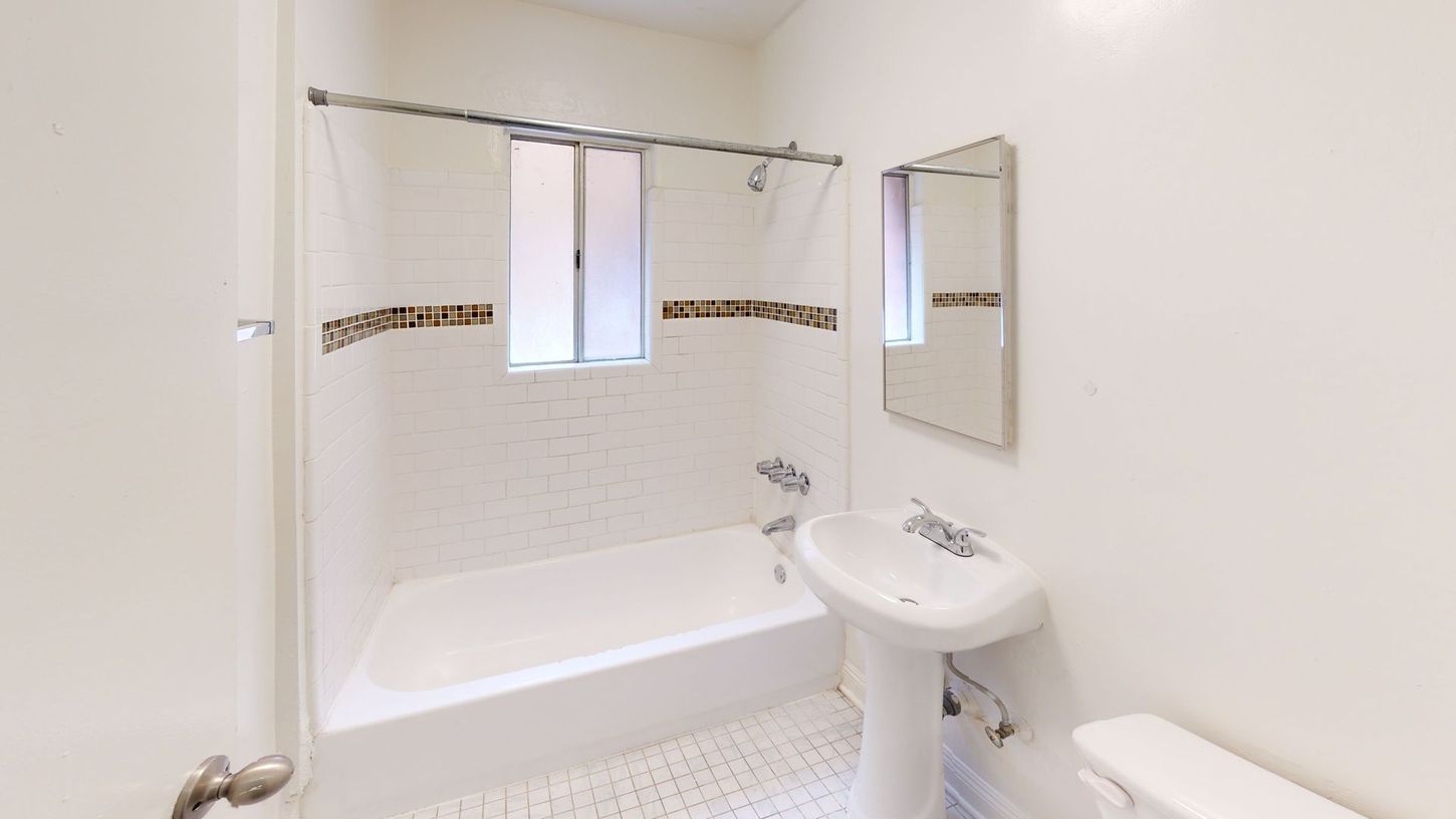 Bathroom with white walls, tub, and pedestal sink; small window and mirror visible.
