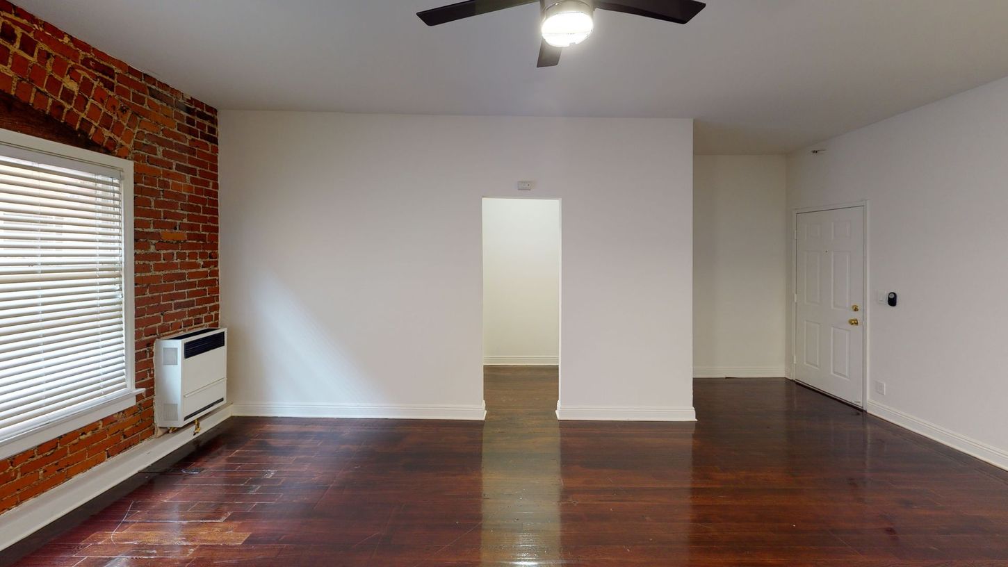 Empty apartment interior with exposed brick wall, wood floors, white walls, and a doorway.