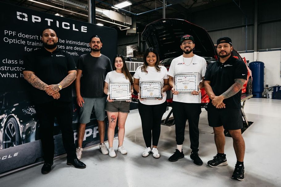 A group of people are standing in front of a car in a garage holding certificates.