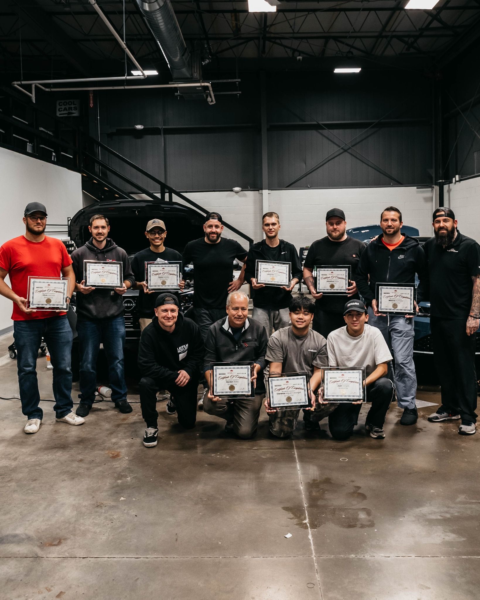 A group of people are standing in front of a car in a garage holding certificates.