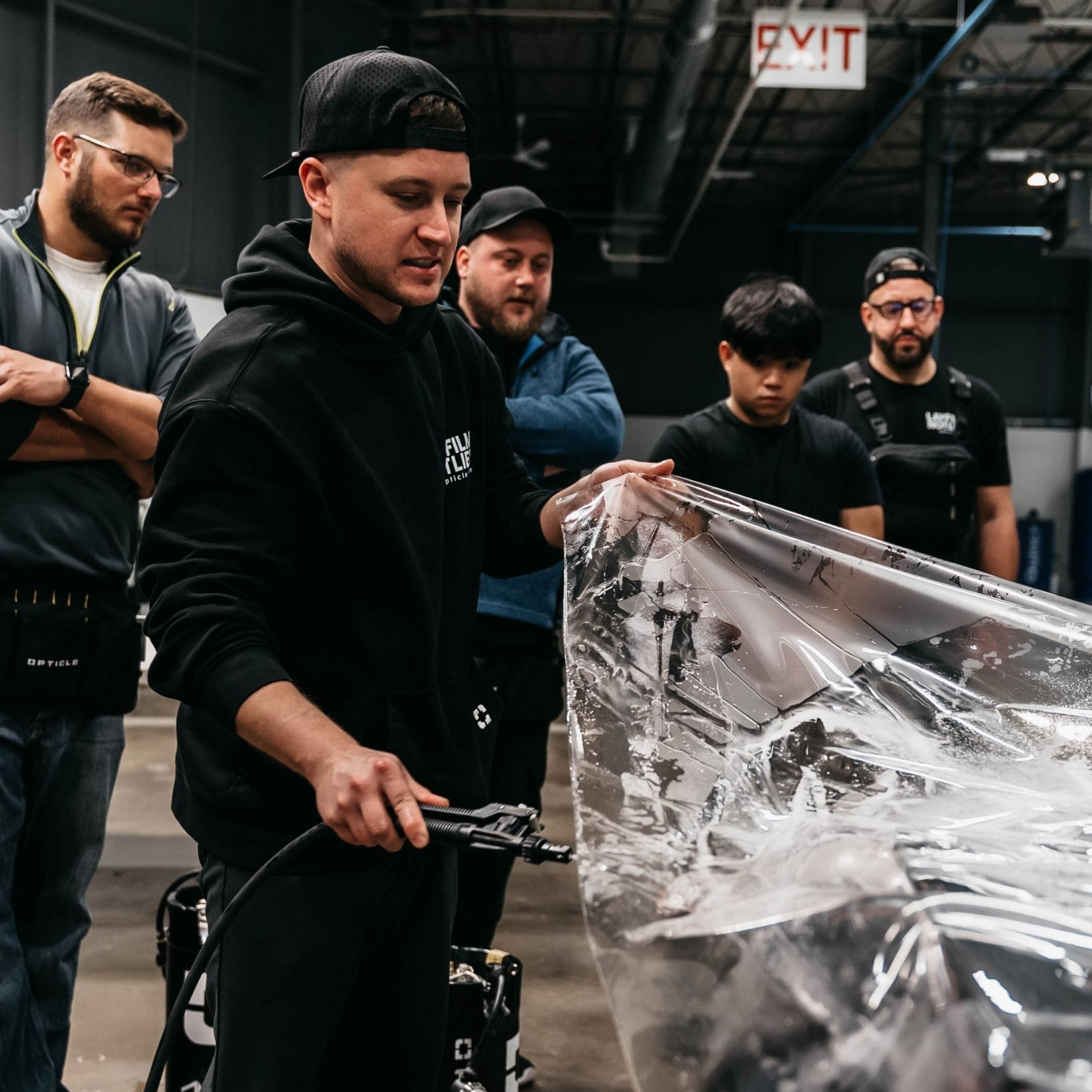 Five people looking at a large, clear sheet, possibly film, being sprayed. Industrial setting.