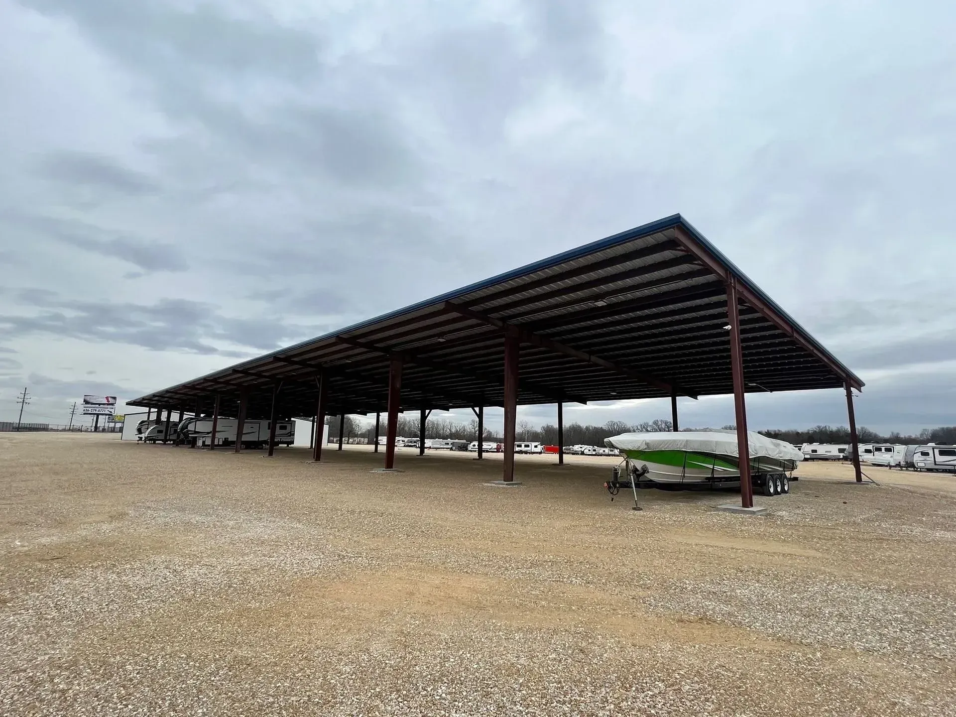 A boat is parked under a covered structure in a gravel lot.