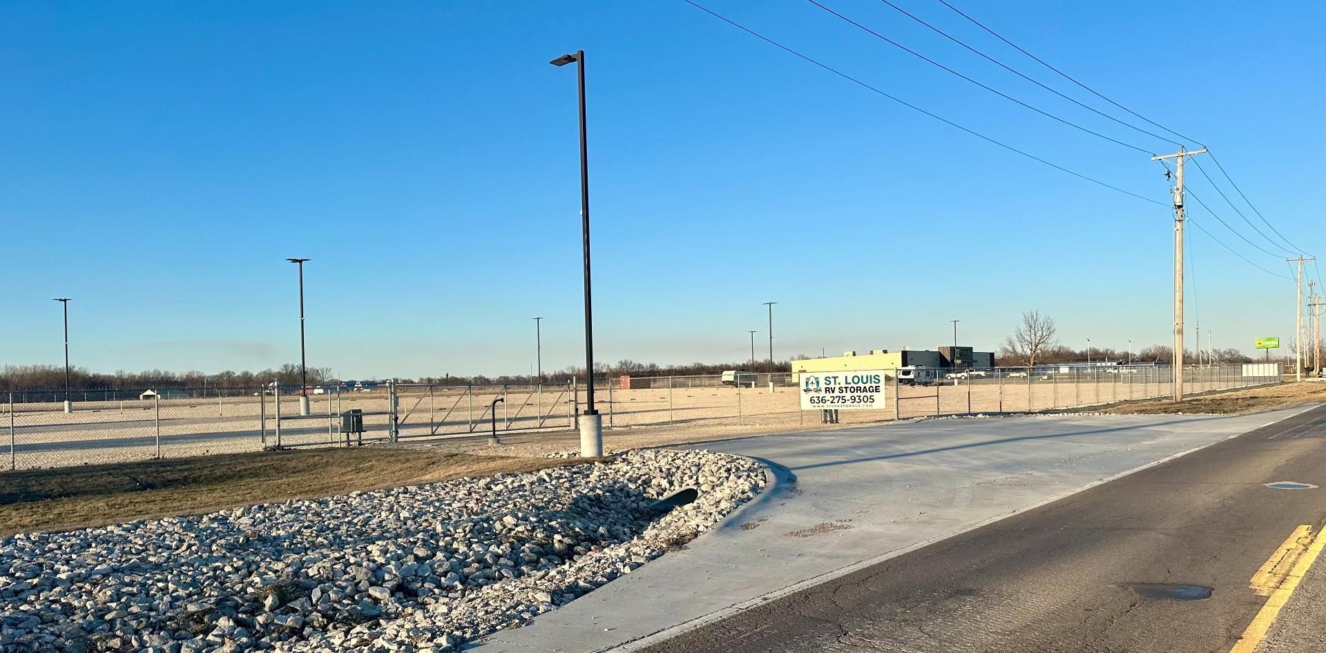 A paved road curves into a large, cleared dirt lot with light poles and a distant building under a clear blue sky.