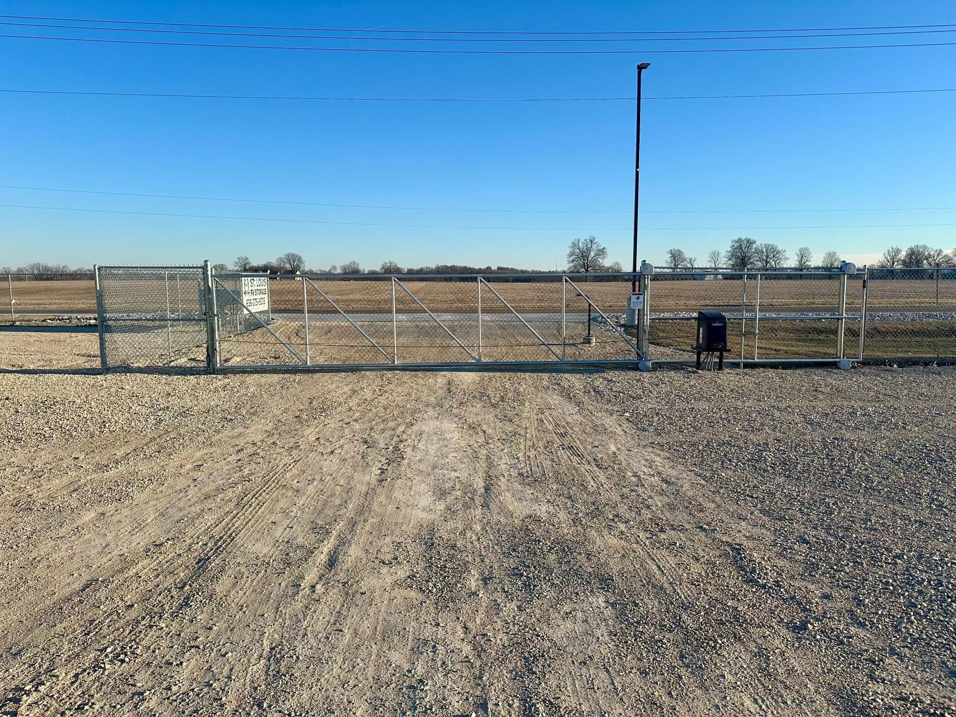 A gravel lot leads to a metal gate and chain-link fence under a clear blue sky, with a security camera mounted on a pole.