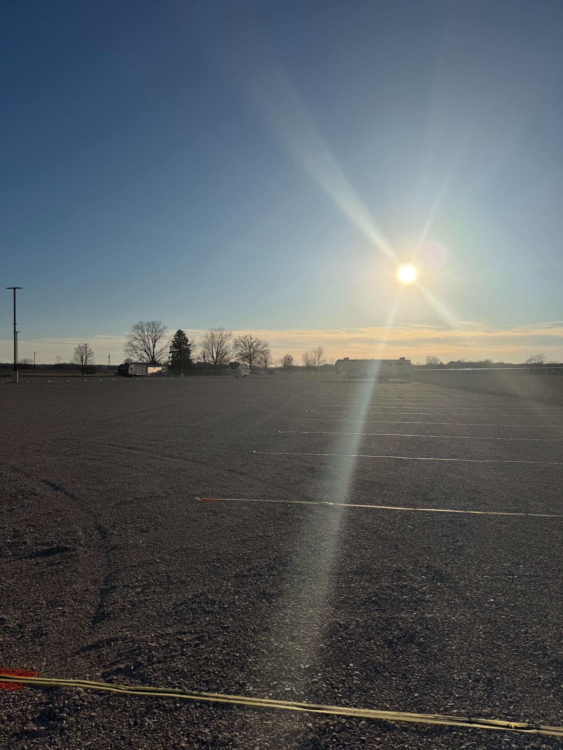 Sun shining over a gravel lot with a few distant trees under a clear blue sky.