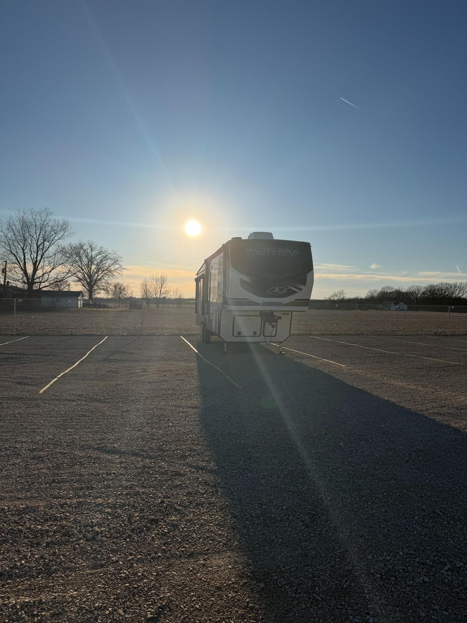 RV parked in a gravel lot with the sun setting behind it, casting a long shadow.