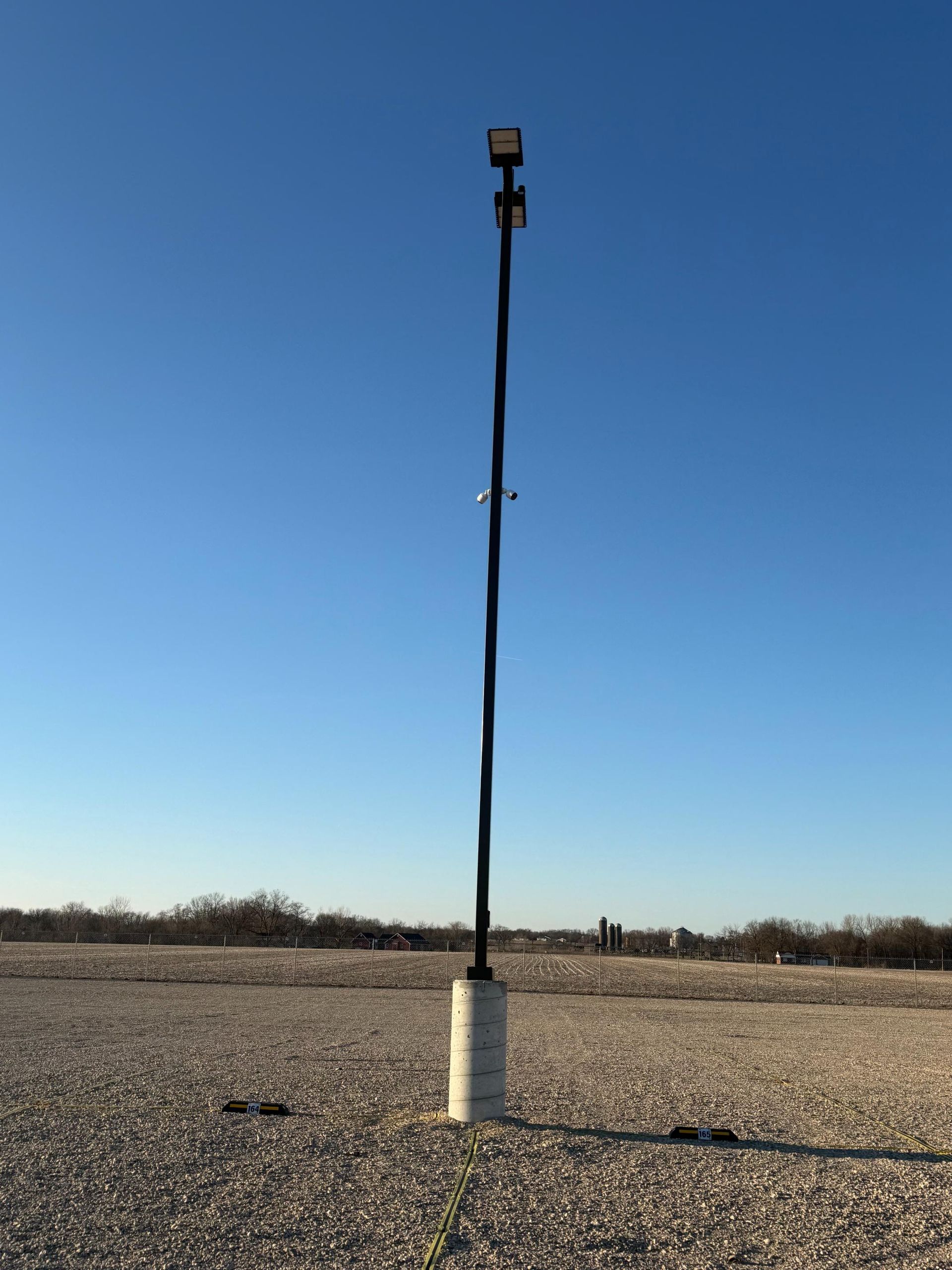A tall black utility pole with lights and cameras on a concrete base against a blue sky.