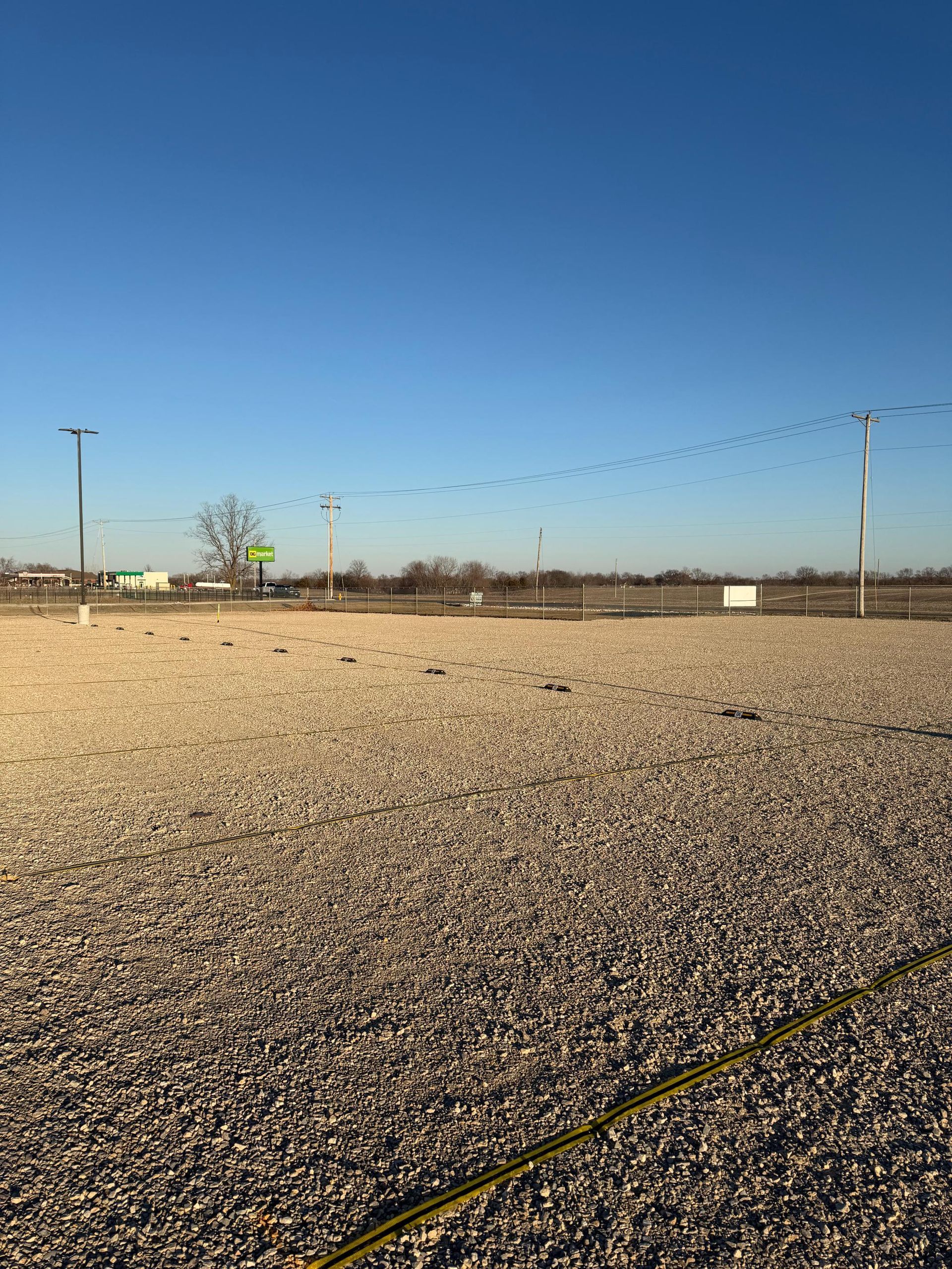 Gravel lot with power lines and birds flying in formation against a clear blue sky.