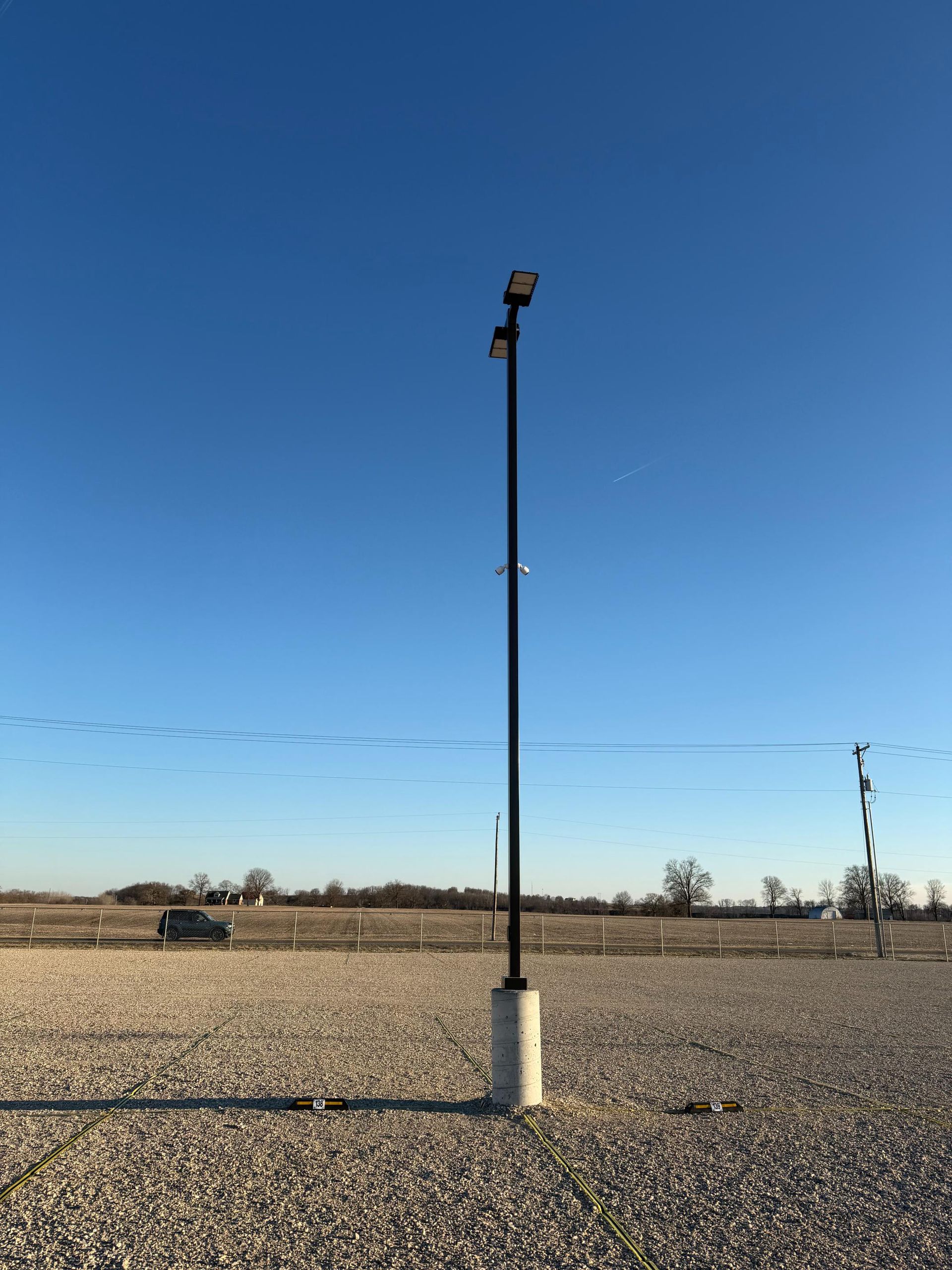 Tall black light pole against a clear blue sky, set in a gravel area with distant trees.