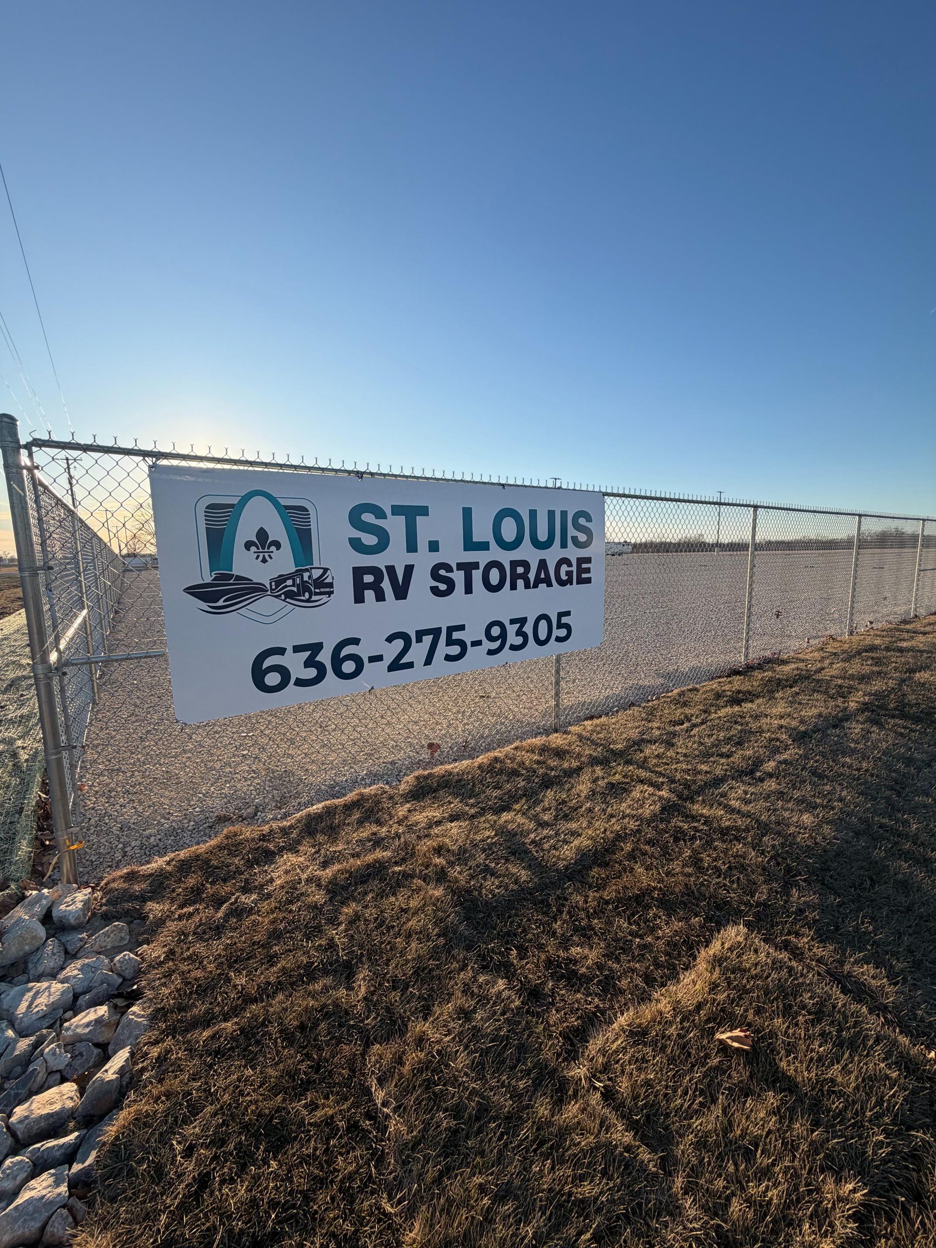 Sign for St. Louis RV Storage on a chain link fence against a sunny, blue sky. Phone number visible.