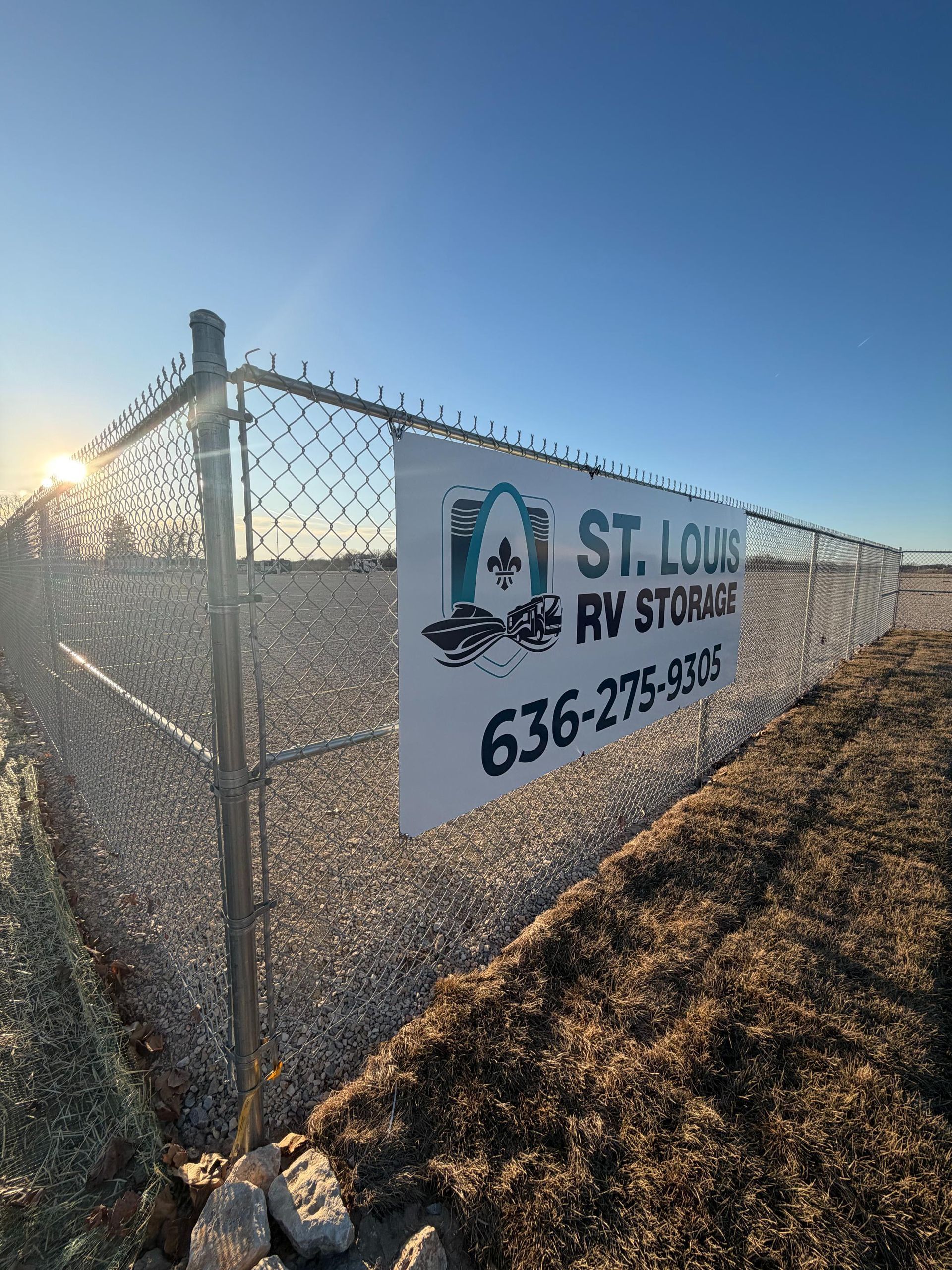 A banner for St. Louis RV Storage with a phone number attached to a chain-link fence at a gravel lot against a blue sky.