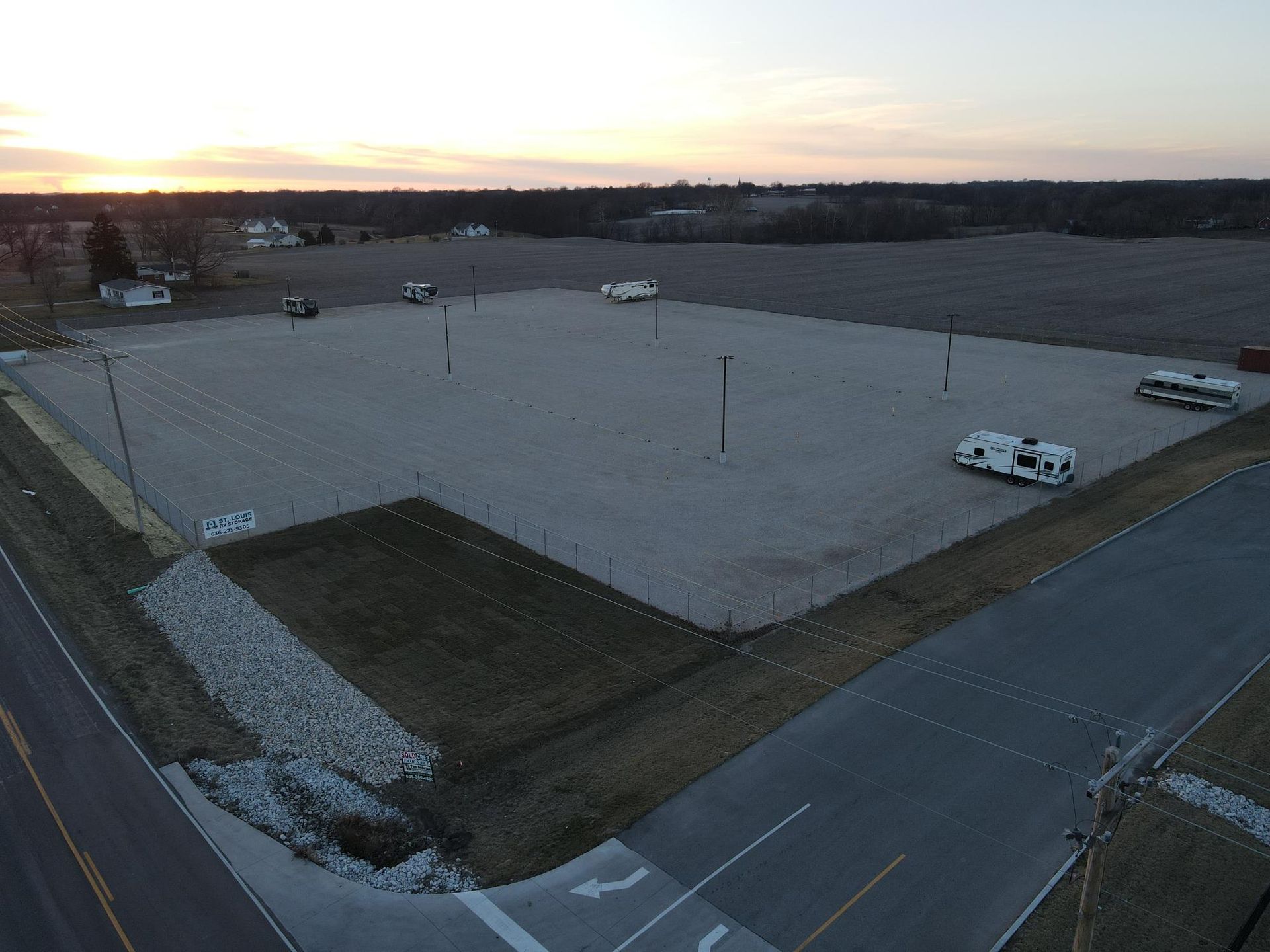 An aerial view of a large, gravel-surfaced parking lot at sunset, featuring a few parked RVs and surrounding fields.
