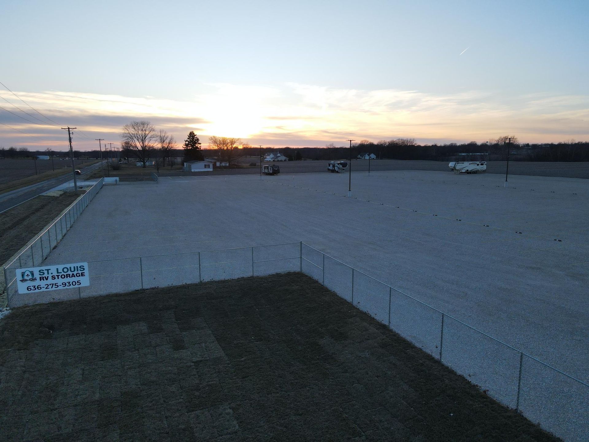 Gravel parking lot at sunset, fenced perimeter, small buildings in background, sign in lower left corner.