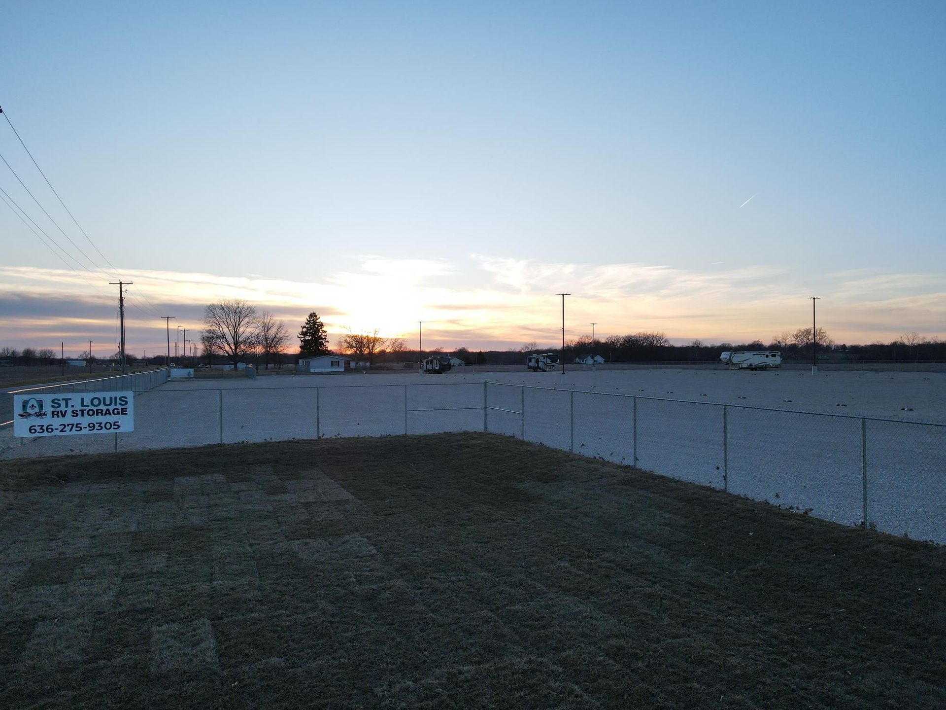 Gravel parking lot at sunset, fenced in, with utility poles, trees, and vehicles; sign in front.