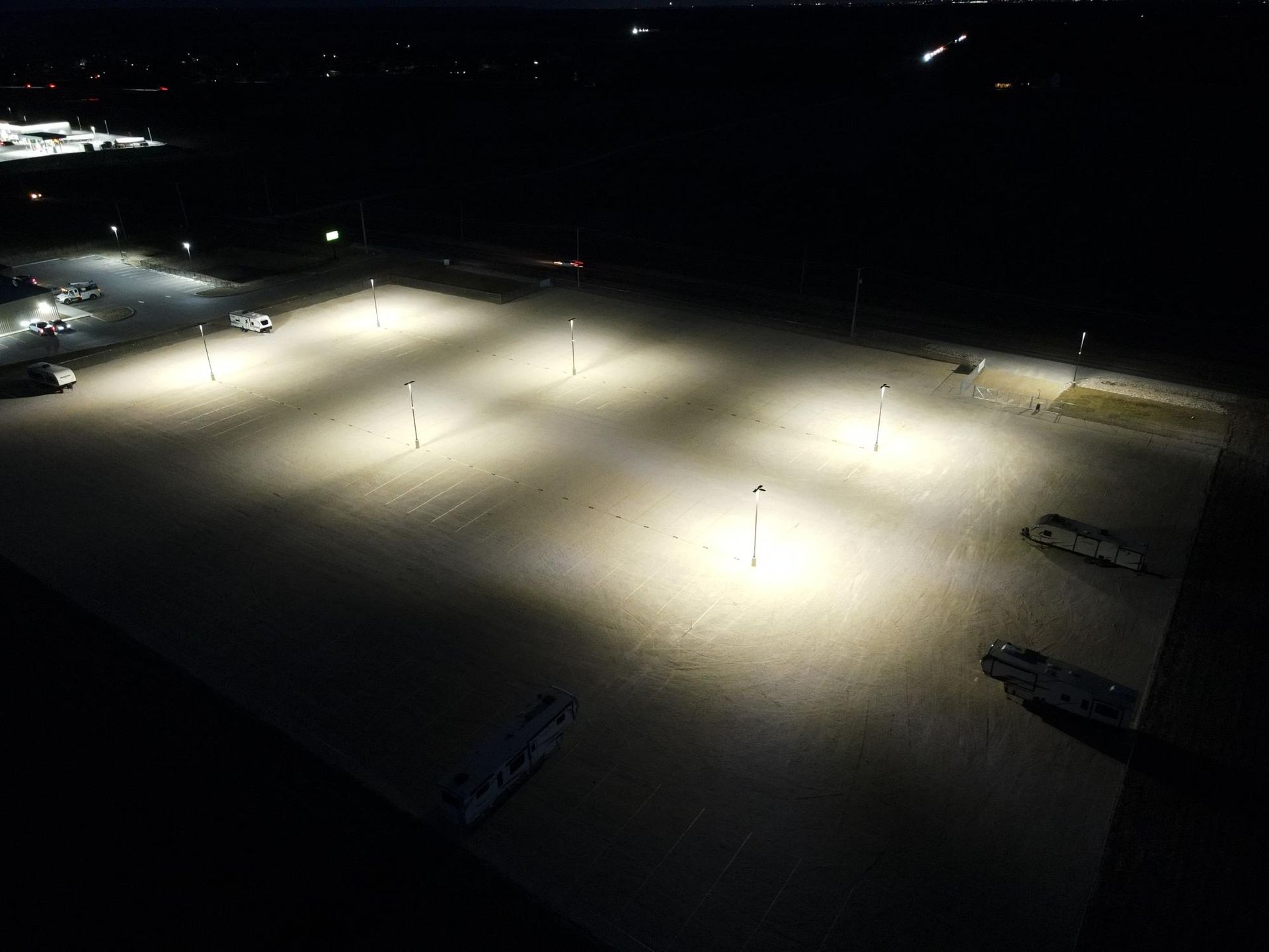 An aerial night view of a parking lot illuminated by tall, bright stadium-style lights, with several parked vehicles.